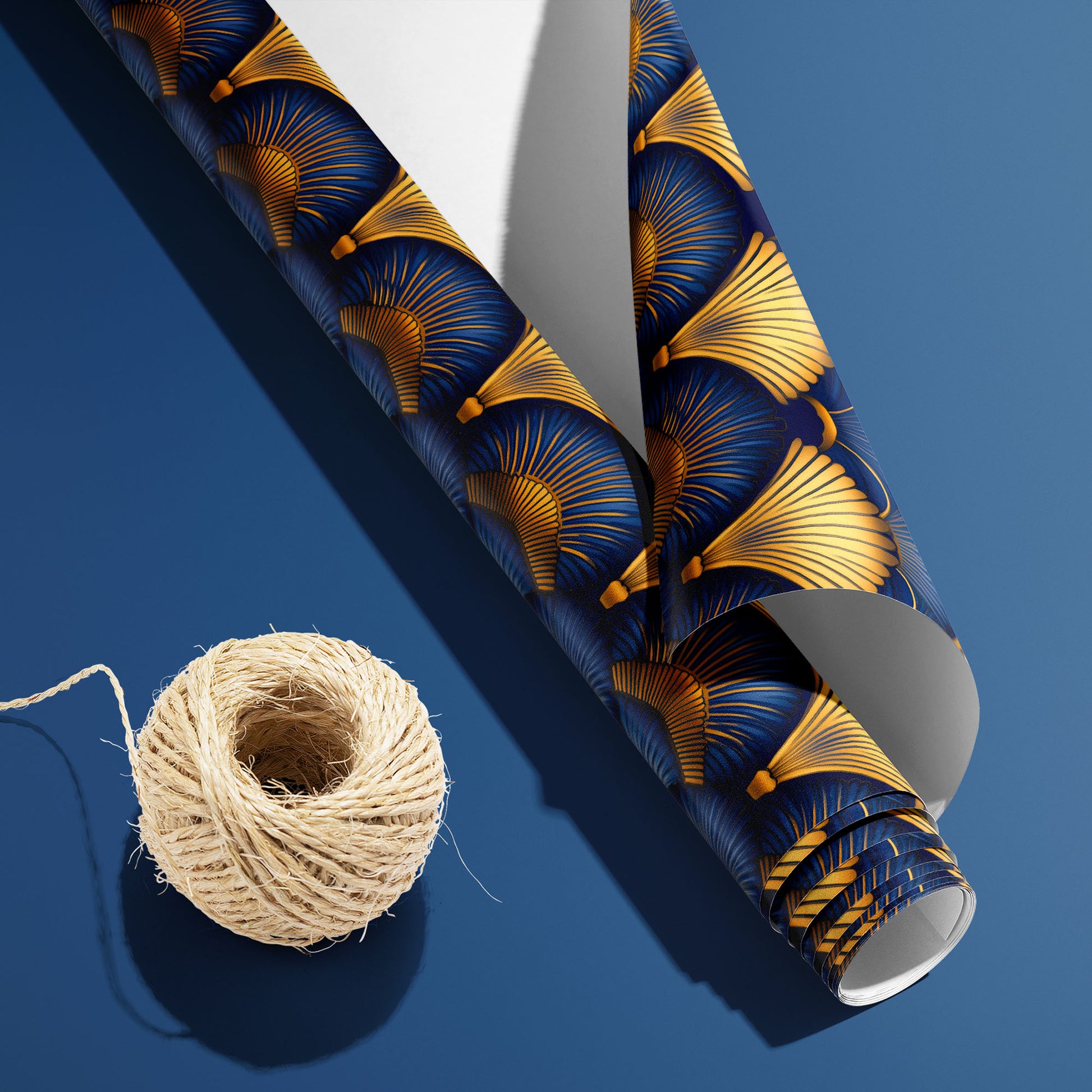A partially unfurled roll of Gilded Ginko with the white underside visible sits ona blue surface beside natural twine, revealing a gold and blue ginkgo leaf pattern.