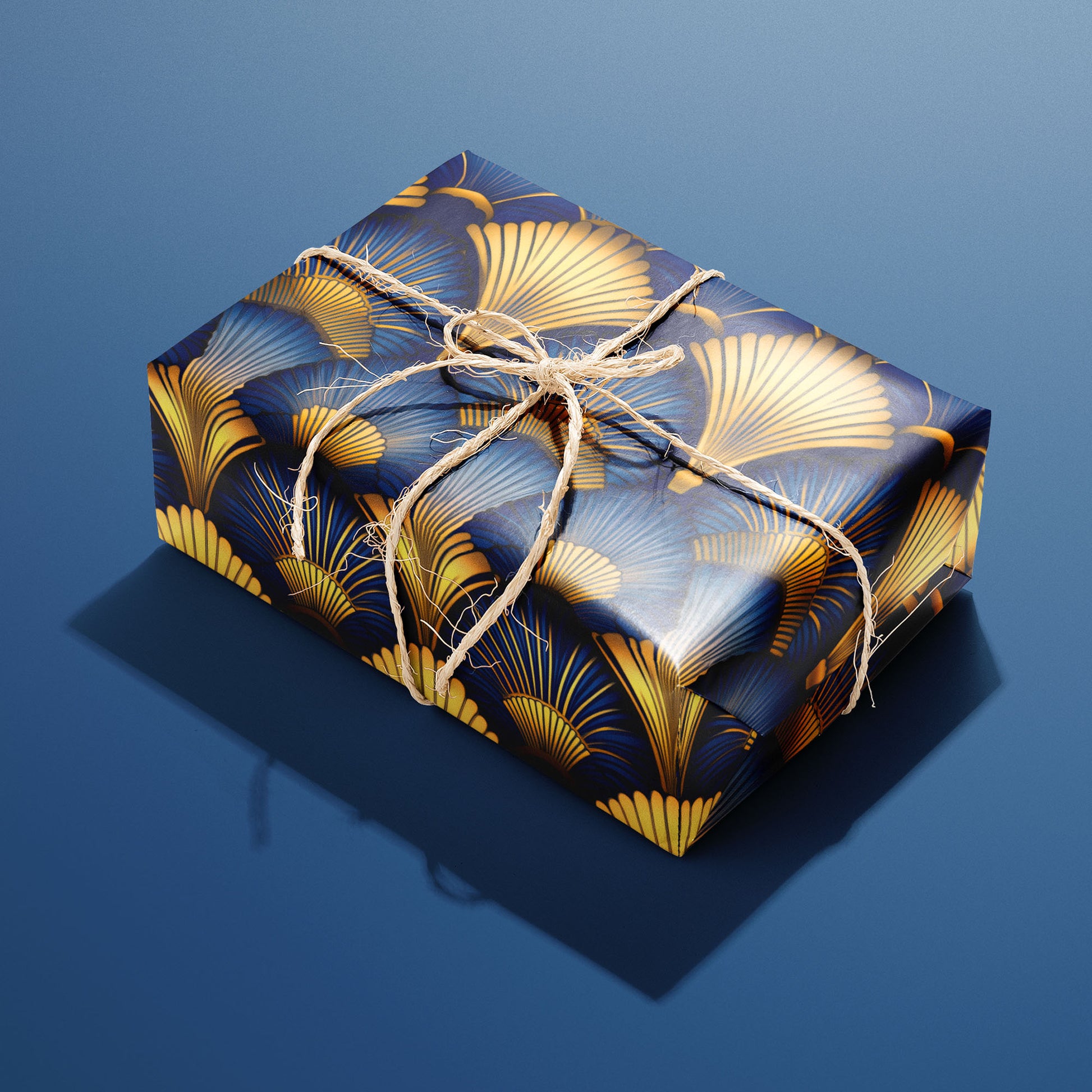A single gift wrapped in Gilded Ginko and tied with natural twine displays gold and blue ginkgo leaves and delicate stems on a dark field, sitting on a blue background.