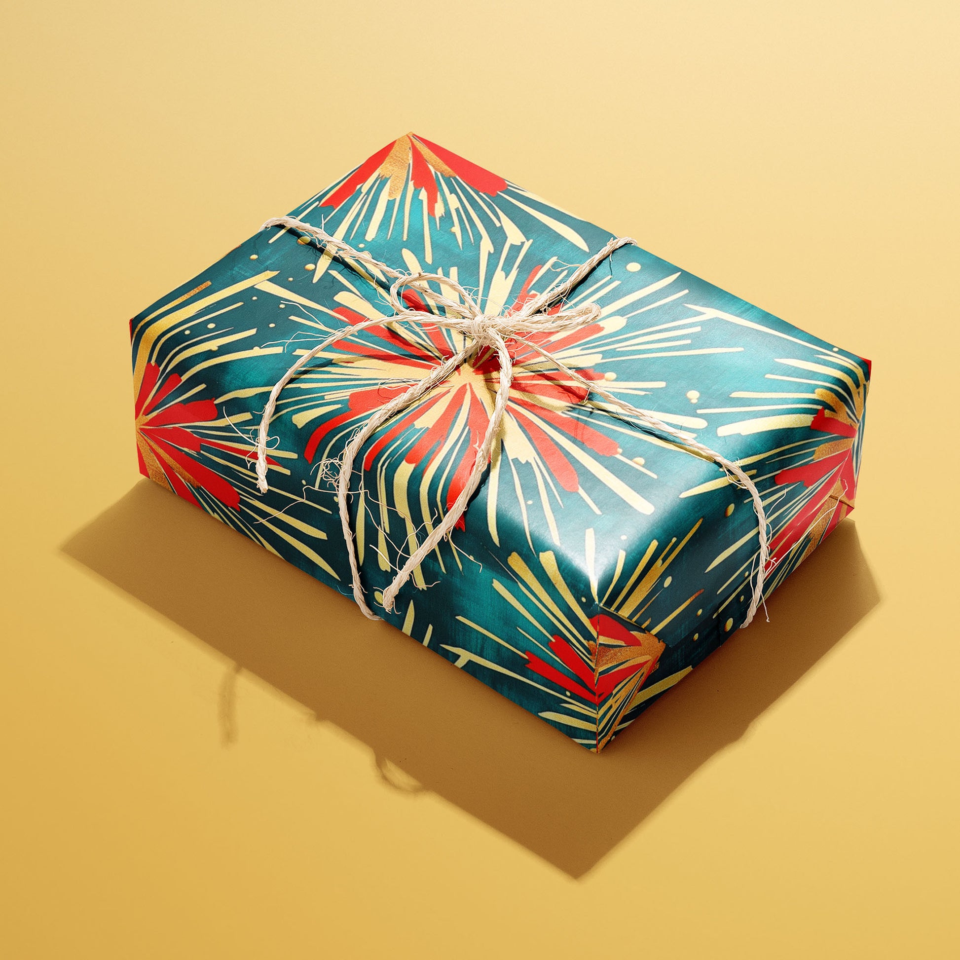 A rectangular gift wrapped in Celestial Celebration paper and tied with natural twine shows red and gold firework bursts on deep teal against a golden yellow background.