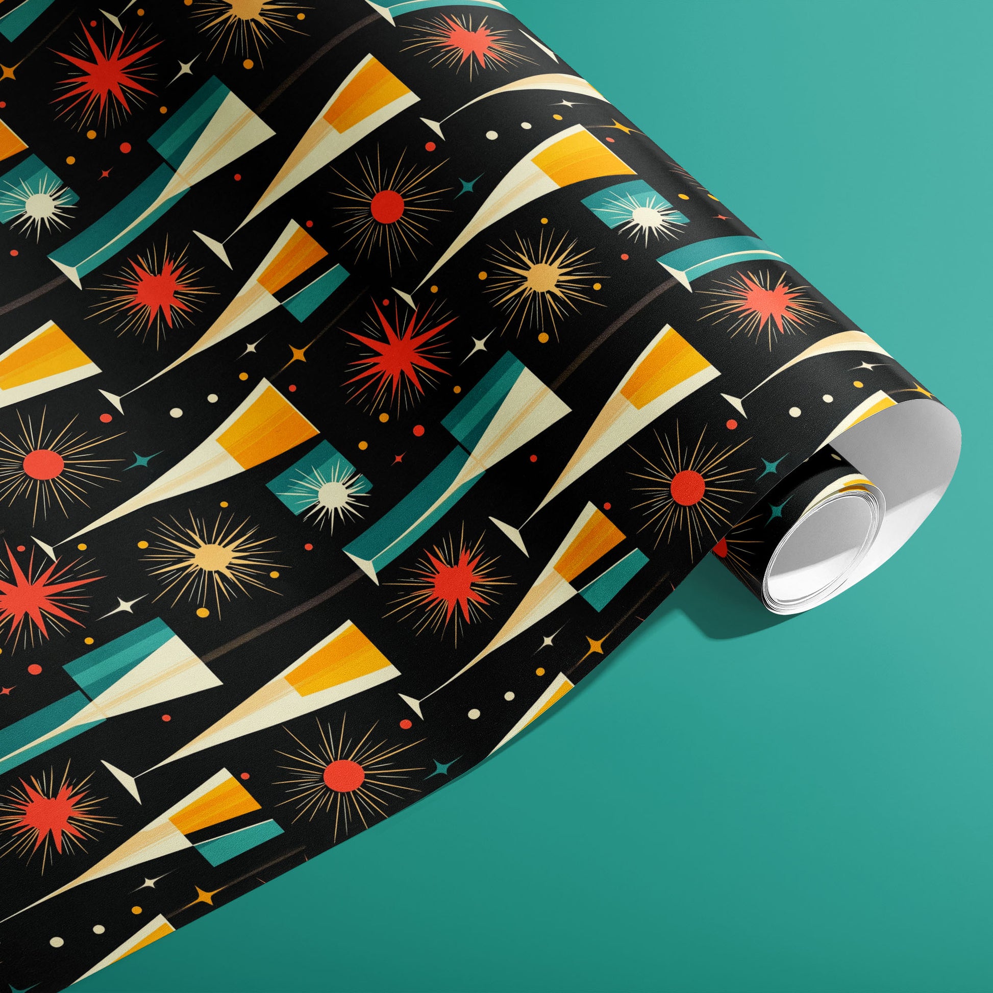 A partially unfurled roll of Midcentury Starburst wrapping paper showing retro starbursts, tapered champagne flutes, and confetti in red, gold, and teal on black, on a teal surface.