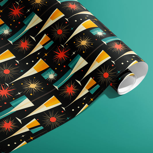 A partially unfurled roll of Midcentury Starburst wrapping paper showing retro starbursts, tapered champagne flutes, and confetti in red, gold, and teal on black, on a teal surface.