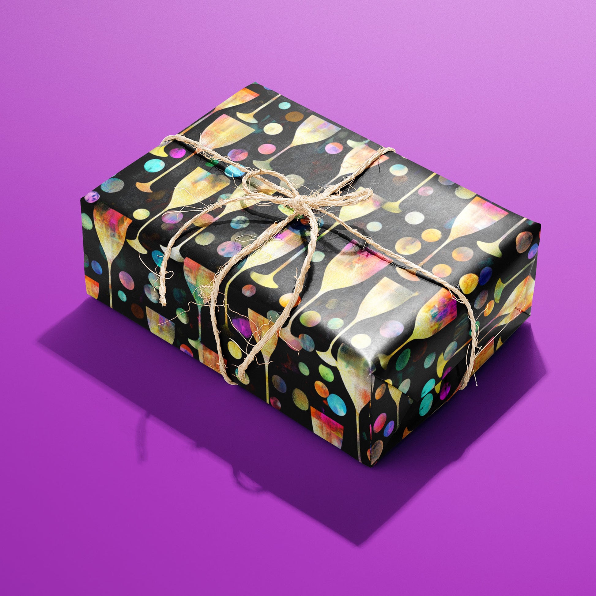 A rectangular gift wrapped in Radiant Revelry paper and tied with natural twine displays glowing champagne flutes and confetti bubbles on black against a vivid purple background.
