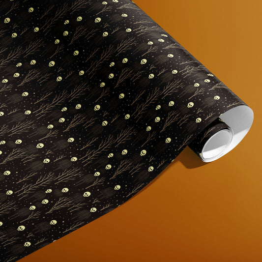 A partially unfurled roll of Skeletons wrapping paper reveals pale skulls and stick-figure skeletons with bare trees and starry specks on black against a warm ochre background.