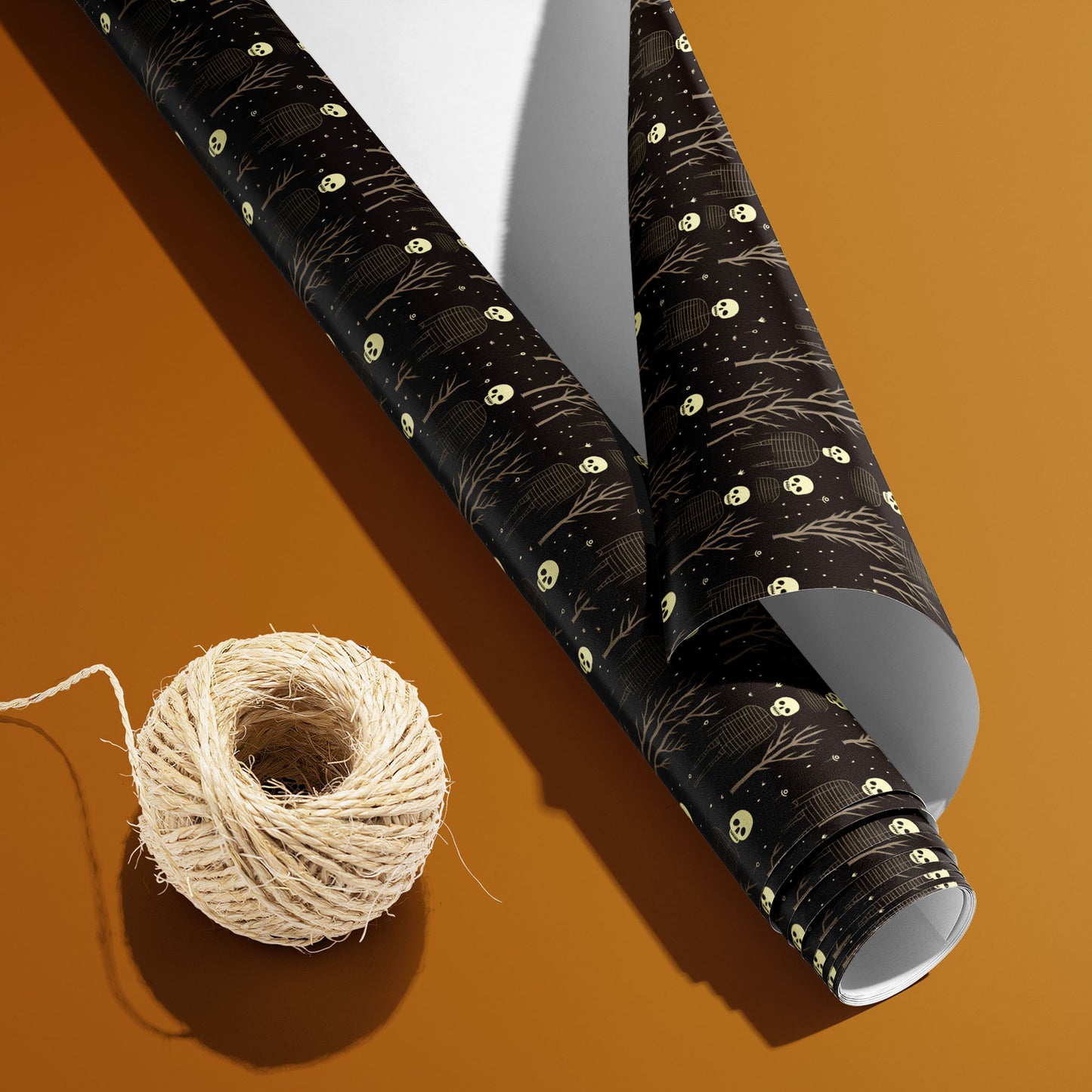 A partially unrolled Skeletons roll with the white underside beside a spool of natural twine displays skulls, ribcage figures, and bare trees on black over a warm ochre background.