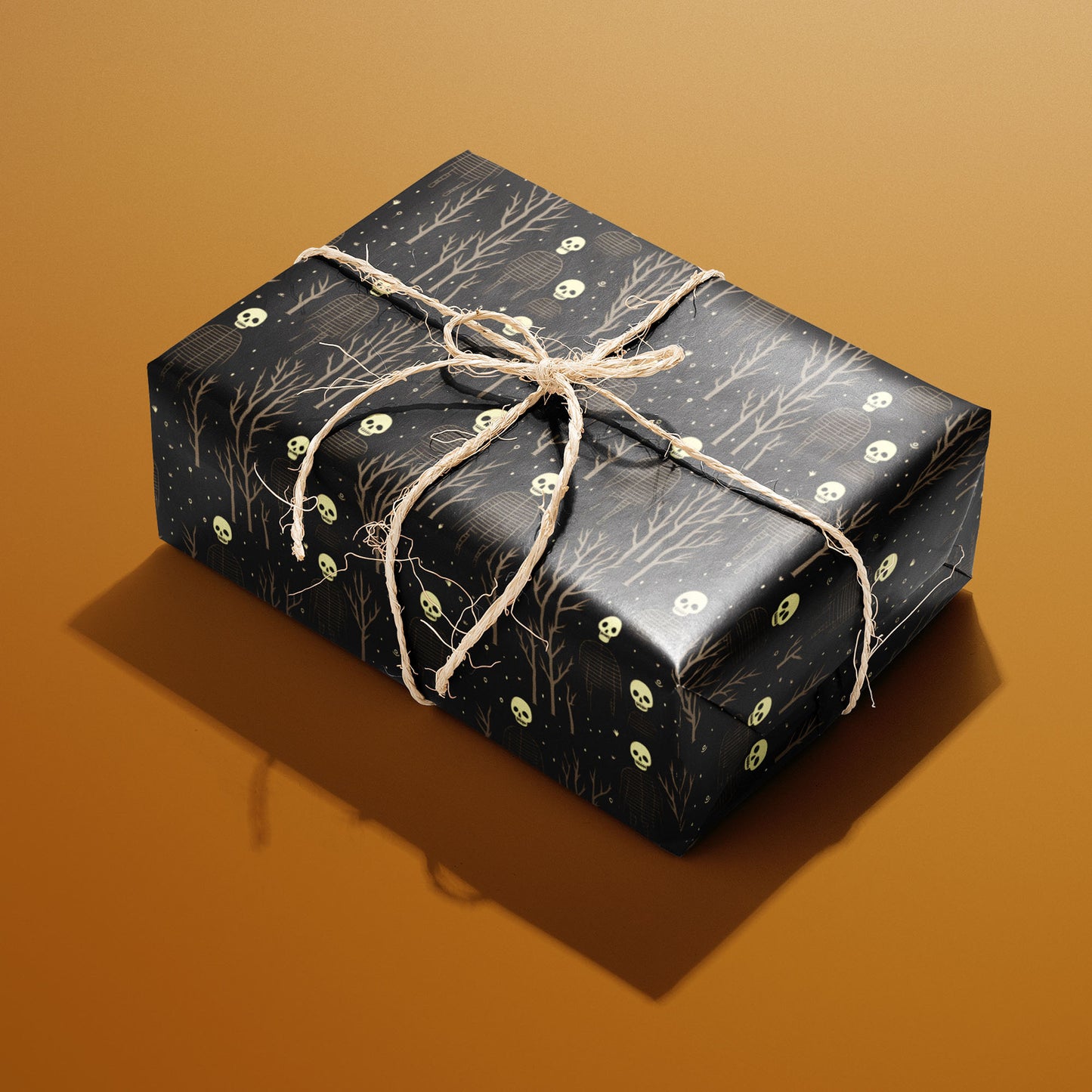 A rectangular gift wrapped in Skeletons paper and tied with natural twine shows skulls and ribcage skeletons amid leafless trees on deep black against a warm ochre background.