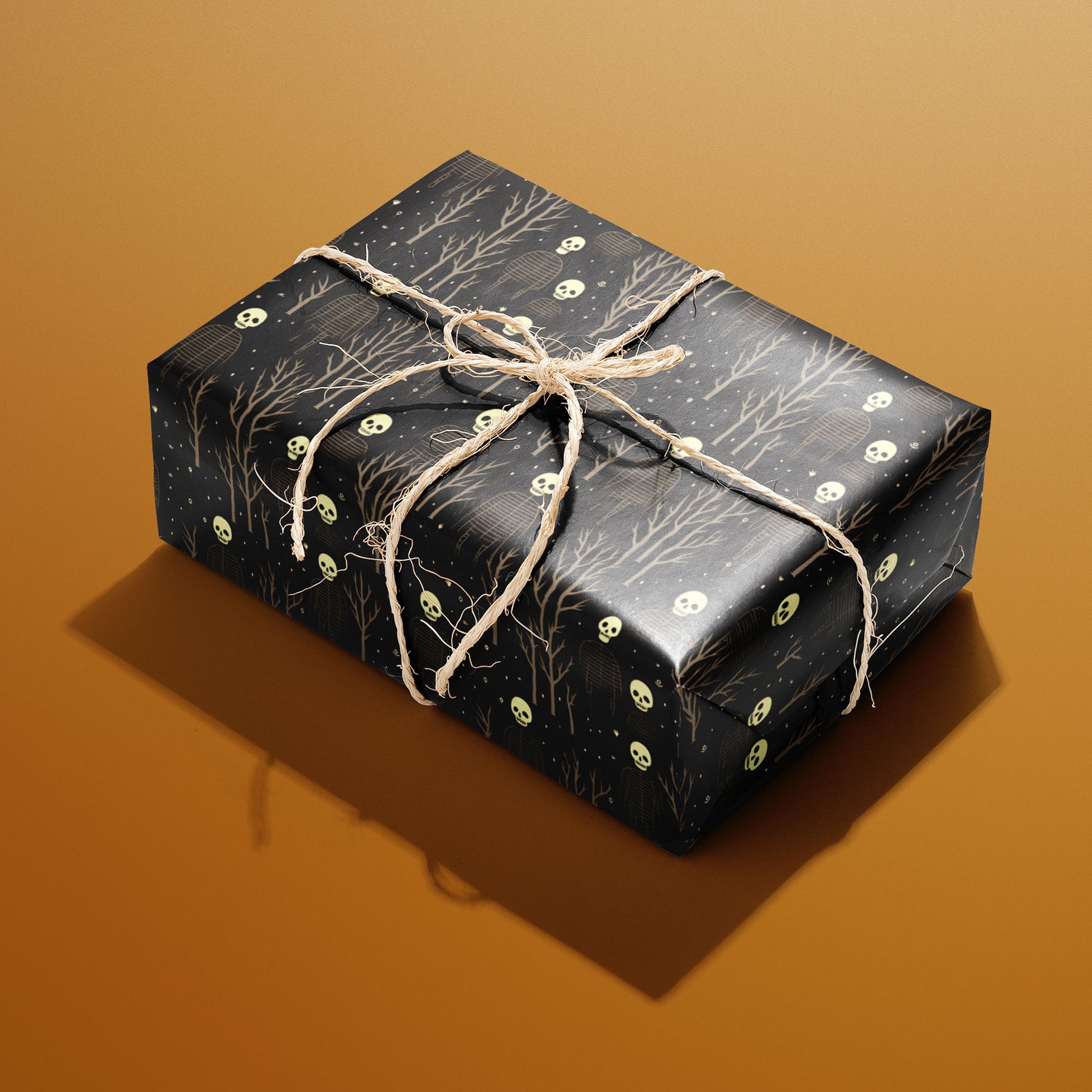 A rectangular gift wrapped in Skeletons paper and tied with natural twine shows skulls and ribcage skeletons amid leafless trees on deep black against a warm ochre background.