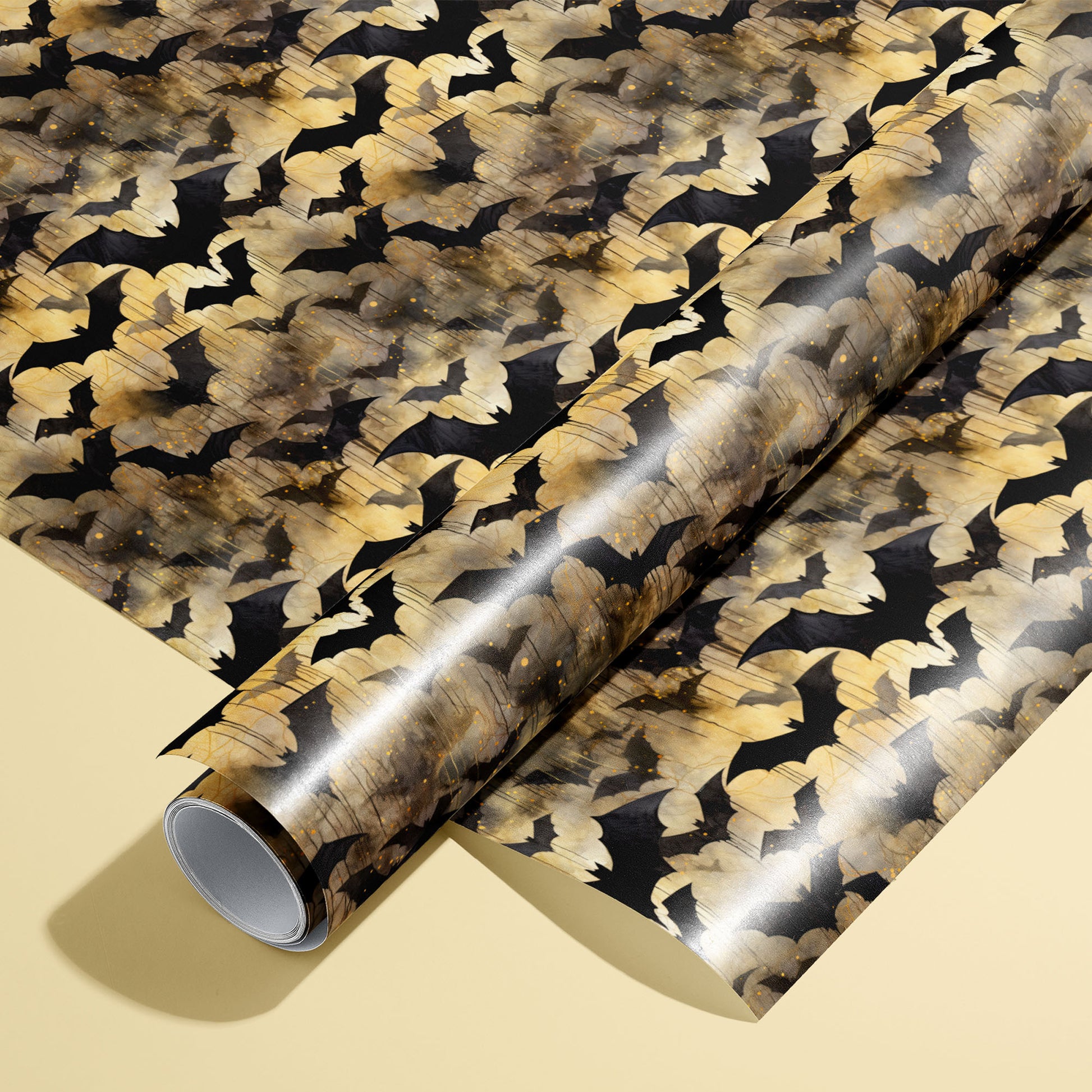 A roll of Spooky Black Bats wrapping paper resting on a matching unrolled sheet, featuring black bat silhouettes over moody gold watercolor textures on a pale yellow background.