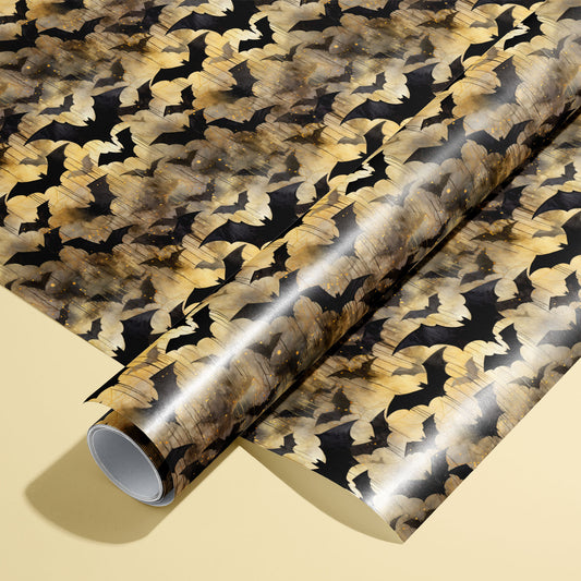 A roll of Spooky Black Bats wrapping paper resting on a matching unrolled sheet, featuring black bat silhouettes over moody gold watercolor textures on a pale yellow background.