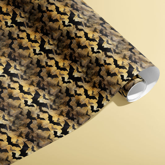 A partially unfurled roll of Spooky Black Bats giftwrap showing inky bat motifs with golden, smoky watercolor washes against a pale yellow surface.