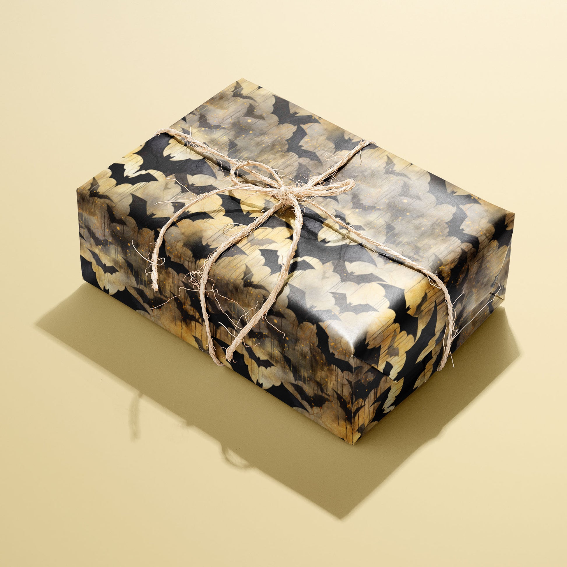 A single rectangular gift wrapped in Spooky Black Bats paper, the bat-and-gold watercolor pattern tied with natural twine, set on a pale yellow background.