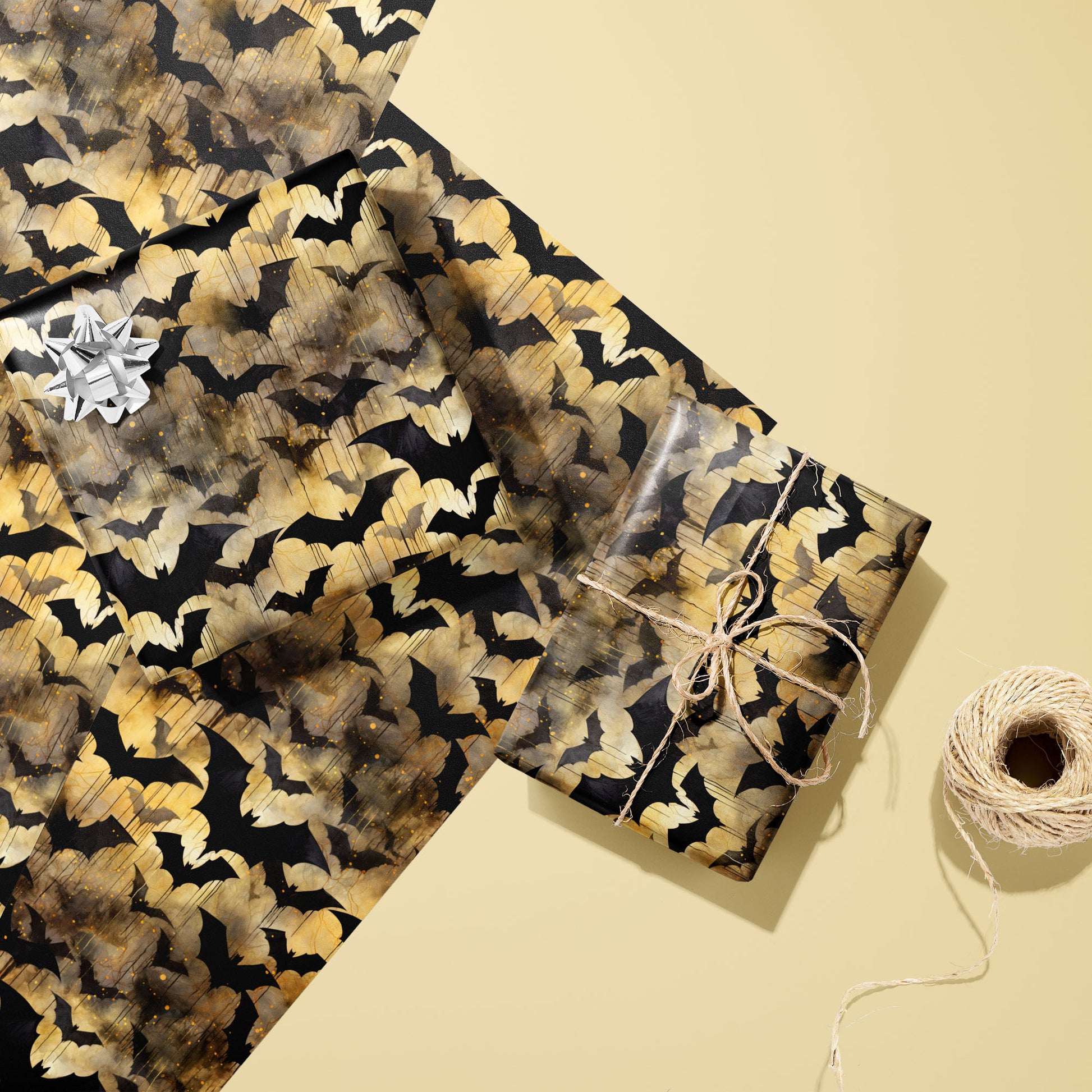 Two gifts wrapped in Spooky Black Bats paper—one topped with a silver bow and one tied with natural twine—rest on two matching sheets beside a twine spool on a pale yellow surface.
