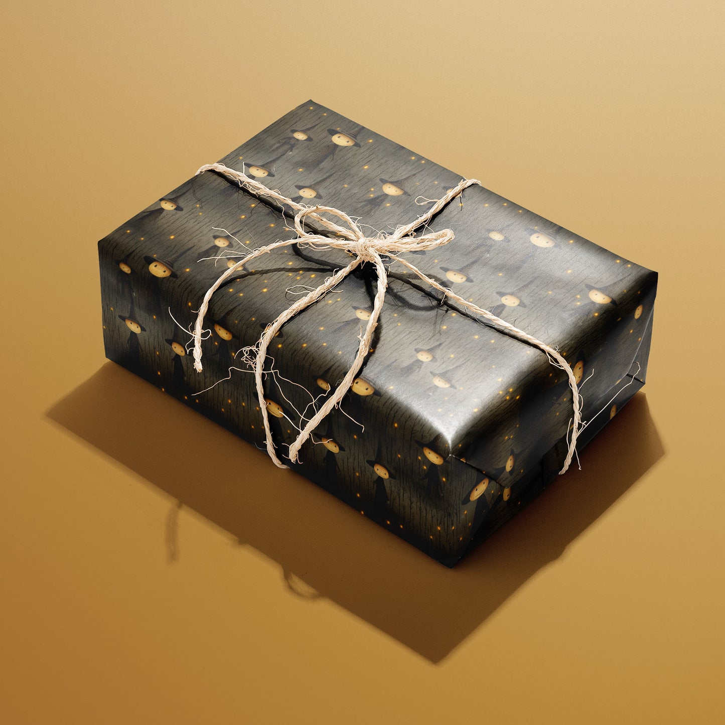 A single rectangular gift wrapped in Halloween Witches paper—pointy-hatted witches and golden specks on charcoal woodgrain—tied with natural twine on a warm mustard background.