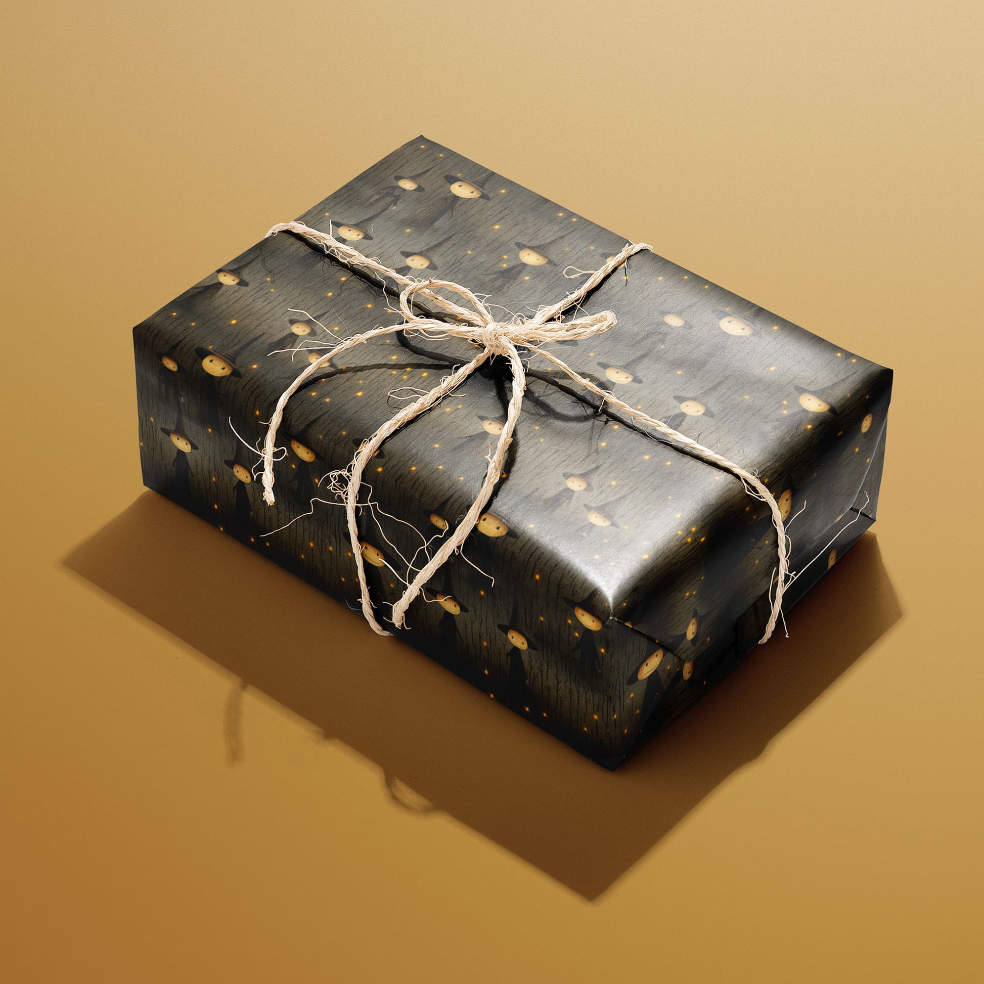 A single rectangular gift wrapped in Halloween Witches paper—pointy-hatted witches and golden specks on charcoal woodgrain—tied with natural twine on a warm mustard background.