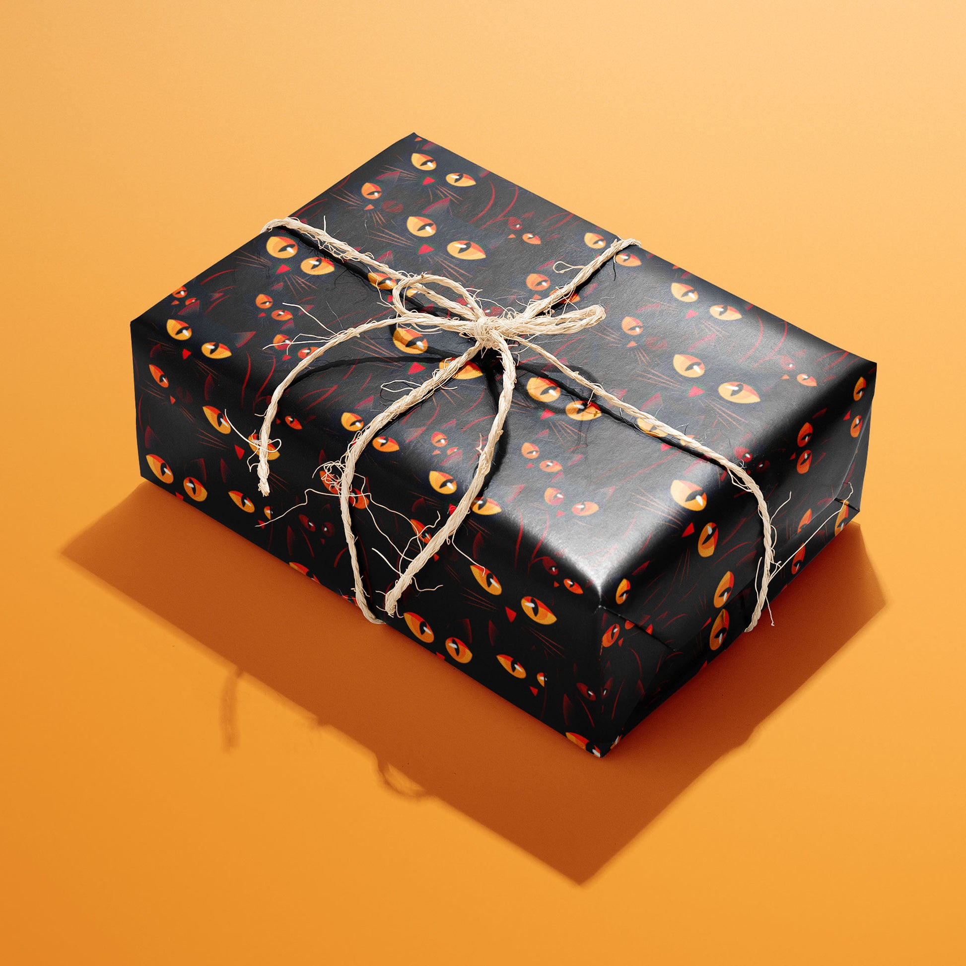 A single rectangular gift wrapped in Cat’s Eyes and tied with natural twine highlights glowing amber eyes and black cat faces on a dark ground against a bright orange backdrop.