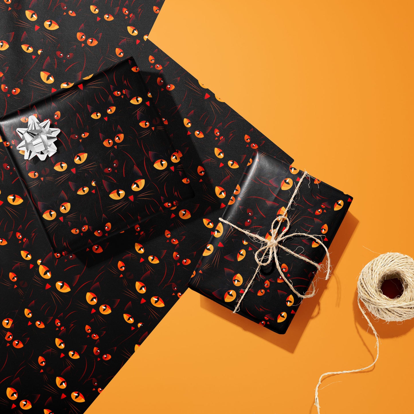 Two Cat’s Eyes gifts—one with a silver bow, one tied with twine—rest on two unused sheets beside a twine spool on bright orange, showing luminous cat eyes on black.