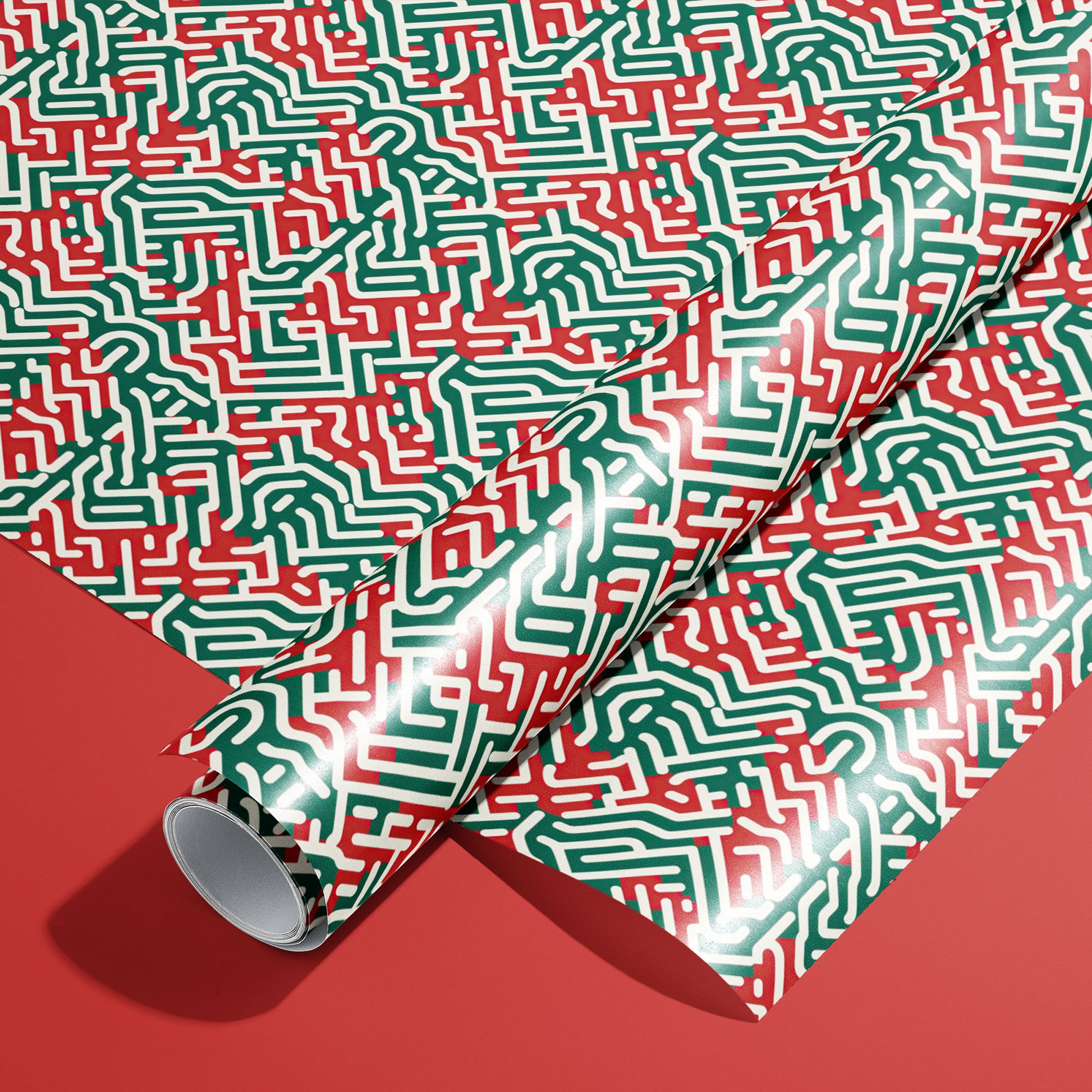 A roll on an unrolled sheet of Geometric Xmas wrapping paper featuring maze-like red, green, and white linework on a satin finish, set on a rich red background.