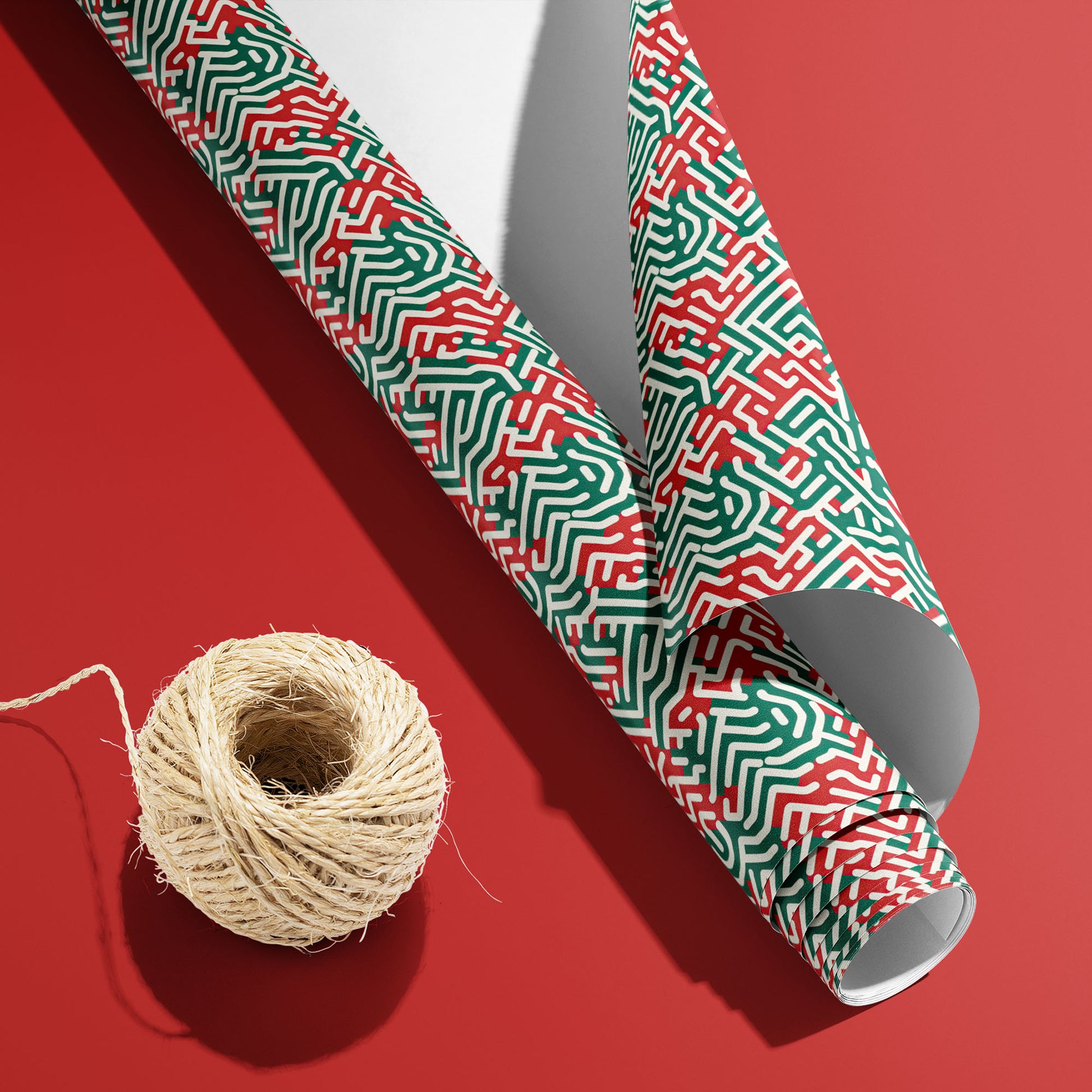 A partially unfurled Geometric Xmas roll with the white underside visible beside a spool of natural twine, showing maze-style holiday linework on a red surface.