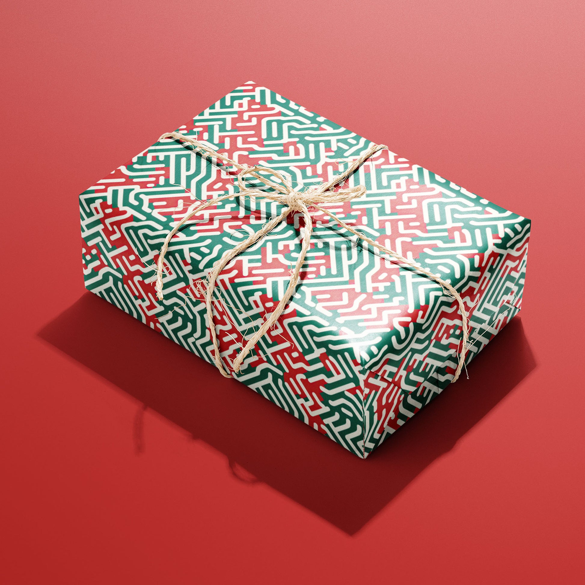 A single rectangular gift wrapped in Geometric Xmas paper tied with natural twine, its red, green, and white geometric maze pattern shining against a red background.