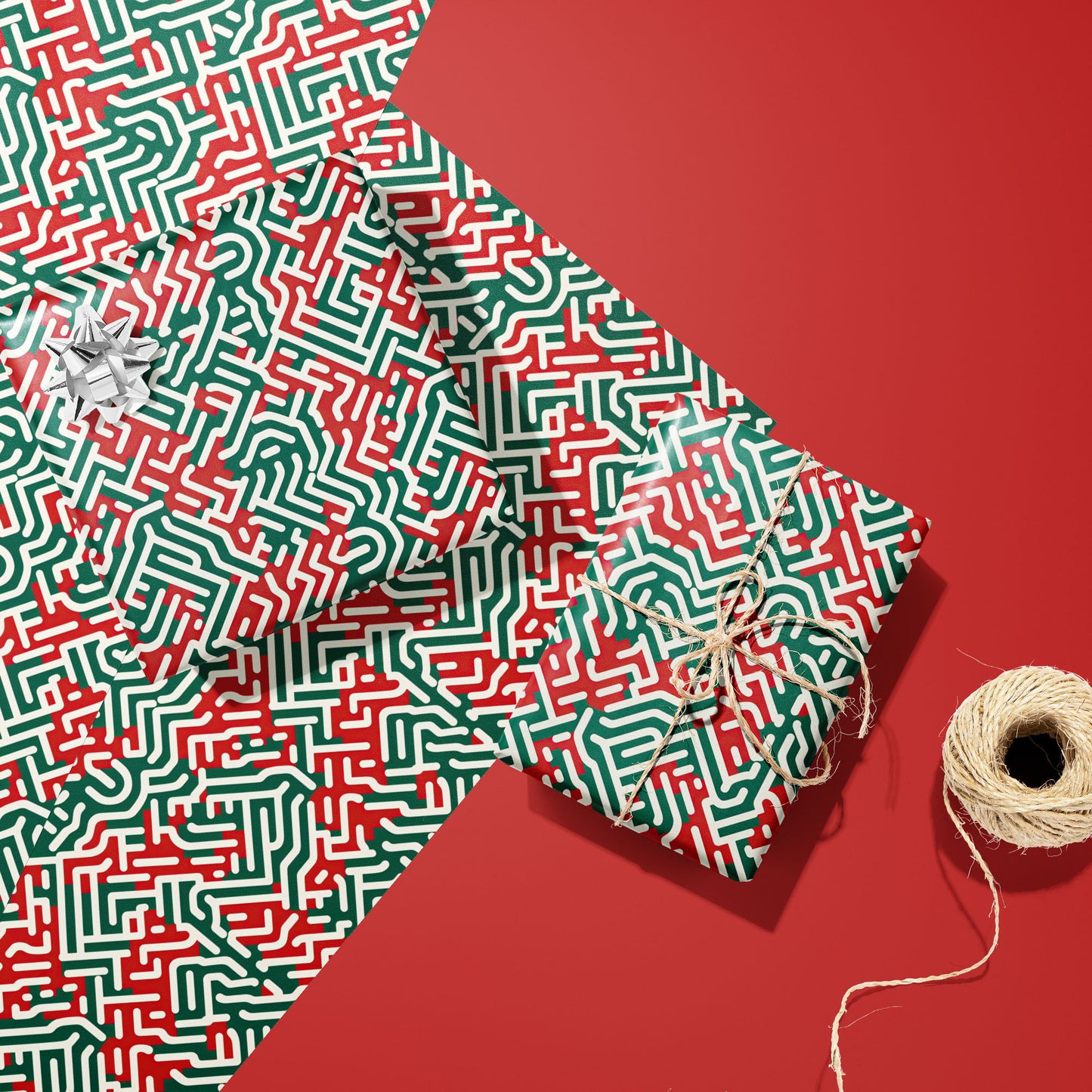 Two gifts in Geometric Xmas wrap—one with a silver bow and one tied with twine—rest on two unused sheets beside a spool of twine on a red background.