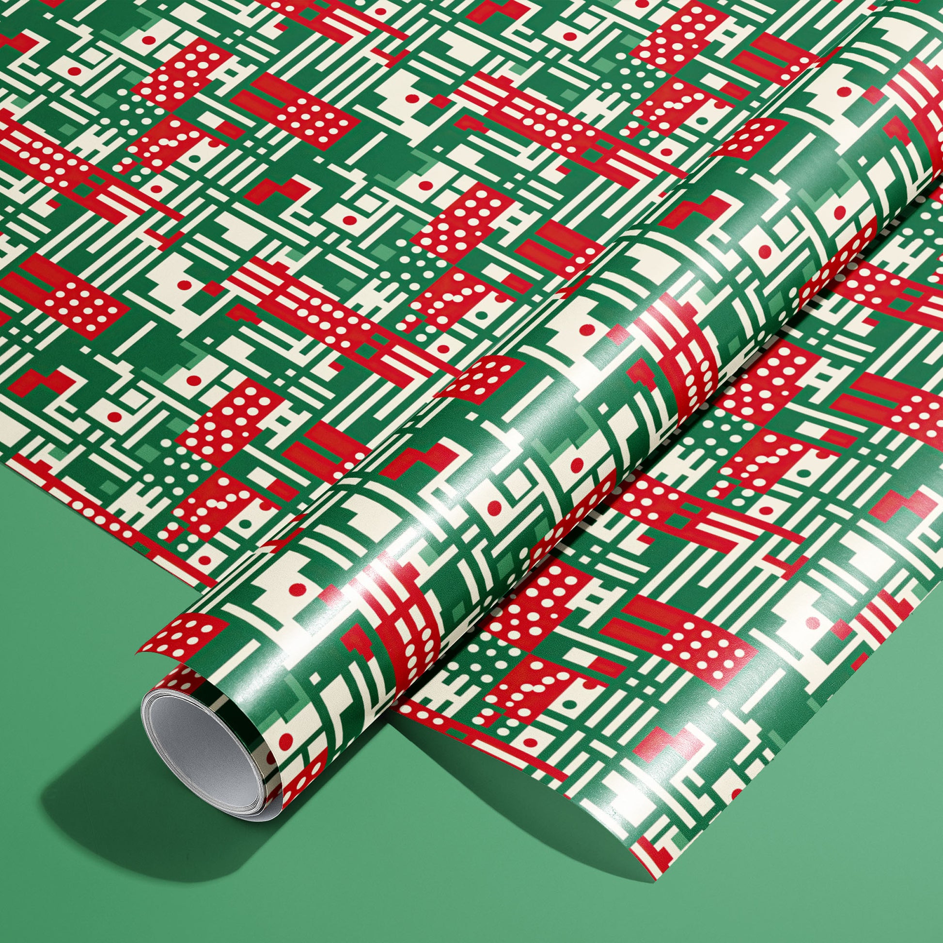 A full roll of Abstract Christmas wrapping paper on an unrolled sheet, showing modern geometric motifs—stripes, dots, and grids—in red, green, and cream on a green background.