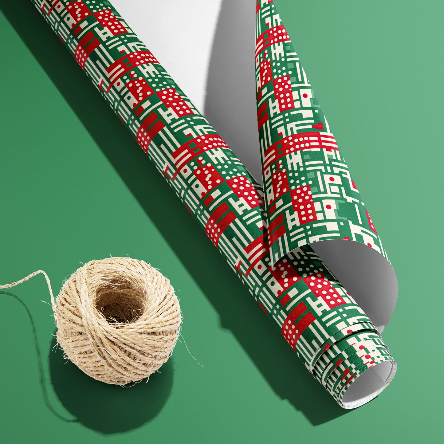 A partially unfurled roll of Abstract Christmas wrapping paper with its white underside visible beside a spool of natural twine on a green background, in red, green, and cream geometric motifs.