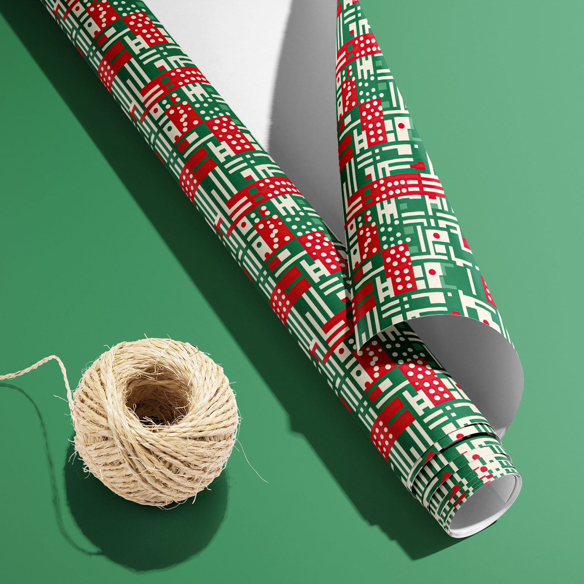 A partially unfurled roll of Abstract Christmas wrapping paper with its white underside visible beside a spool of natural twine on a green background, in red, green, and cream geometric motifs.