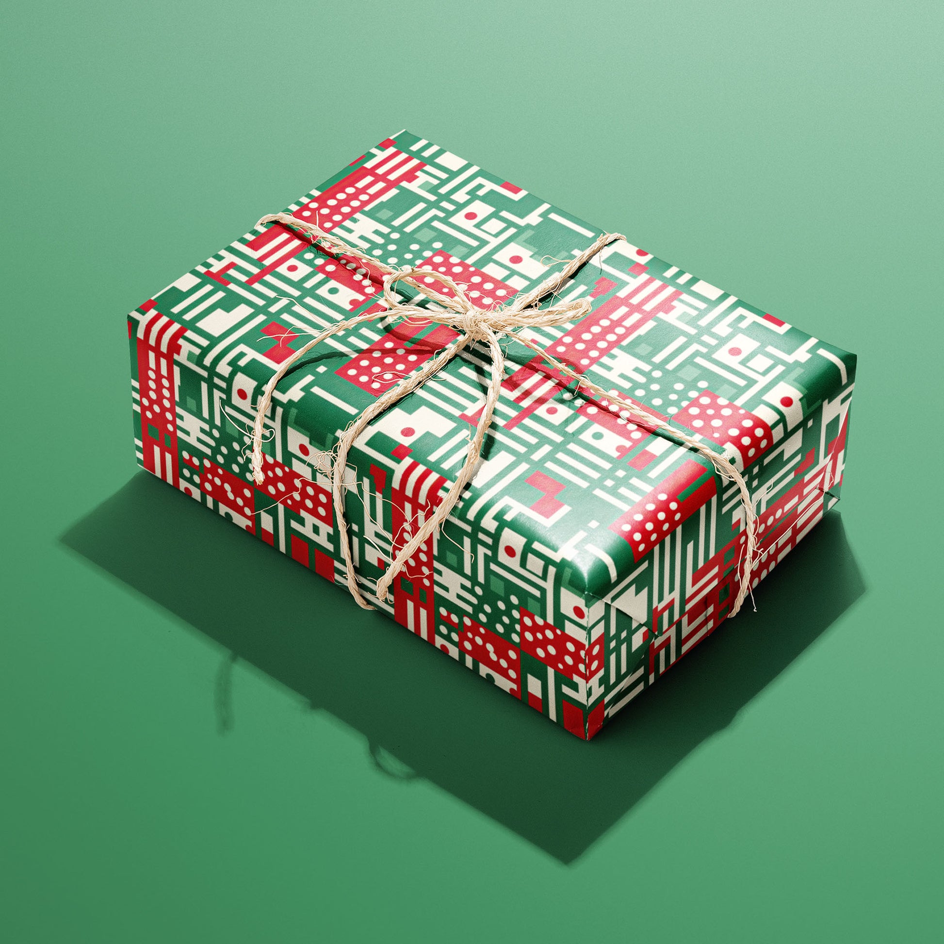 A single gift wrapped in Abstract Christmas paper and tied with natural twine, displaying red, green, and cream geometric stripes, dots, and grids on a green background.