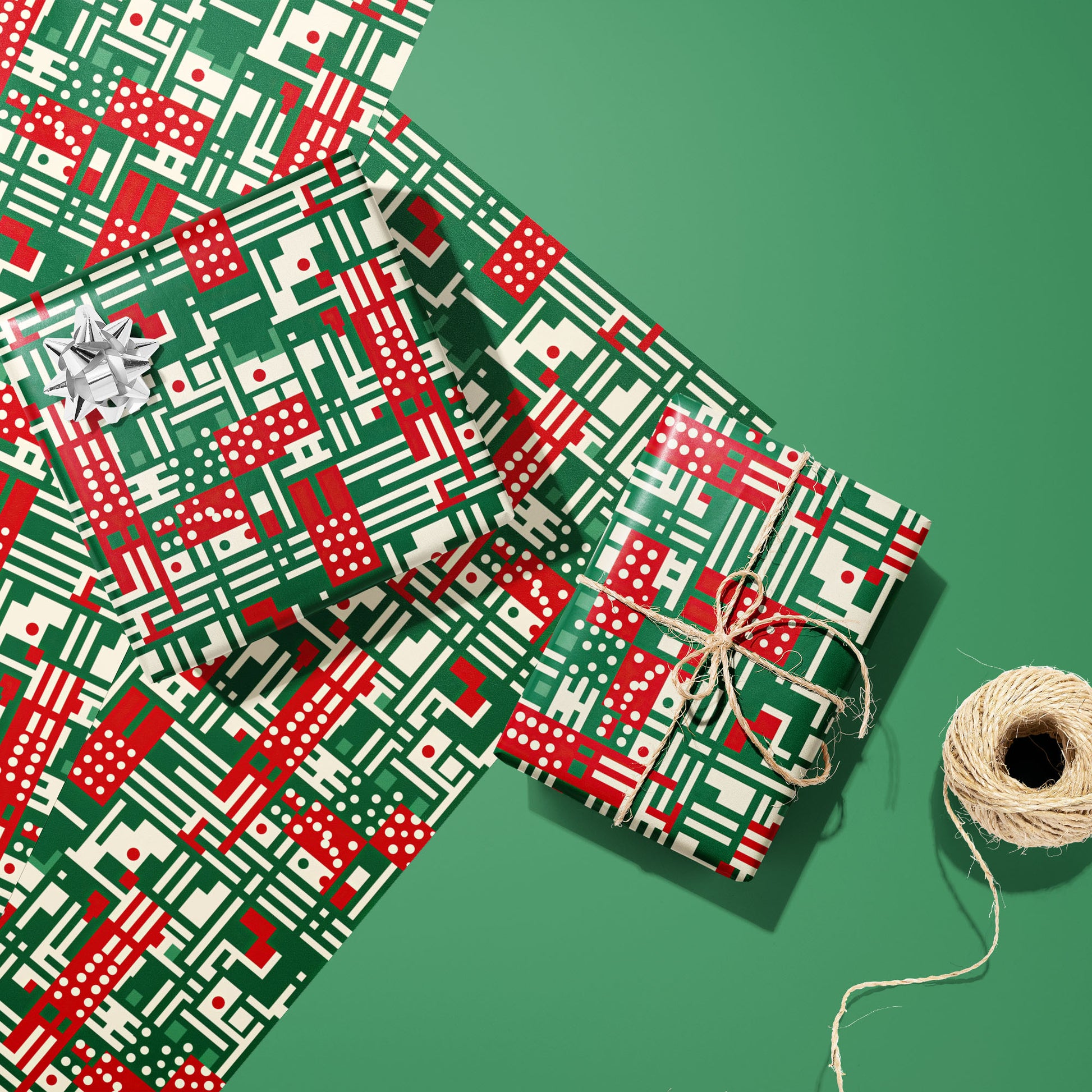 Two gifts wrapped in Abstract Christmas paper—one with a silver bow, one tied with twine—on two unused sheets beside a twine spool on a green background, in red, green, and cream geometric motifs.