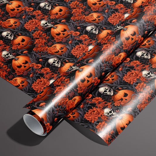 A roll of Ghastly Grove wrapping paper resting on an unrolled sheet, showing jack-o’-lanterns, skulls, and red florals amid dark foliage on a black ground, on a dark gray surface.