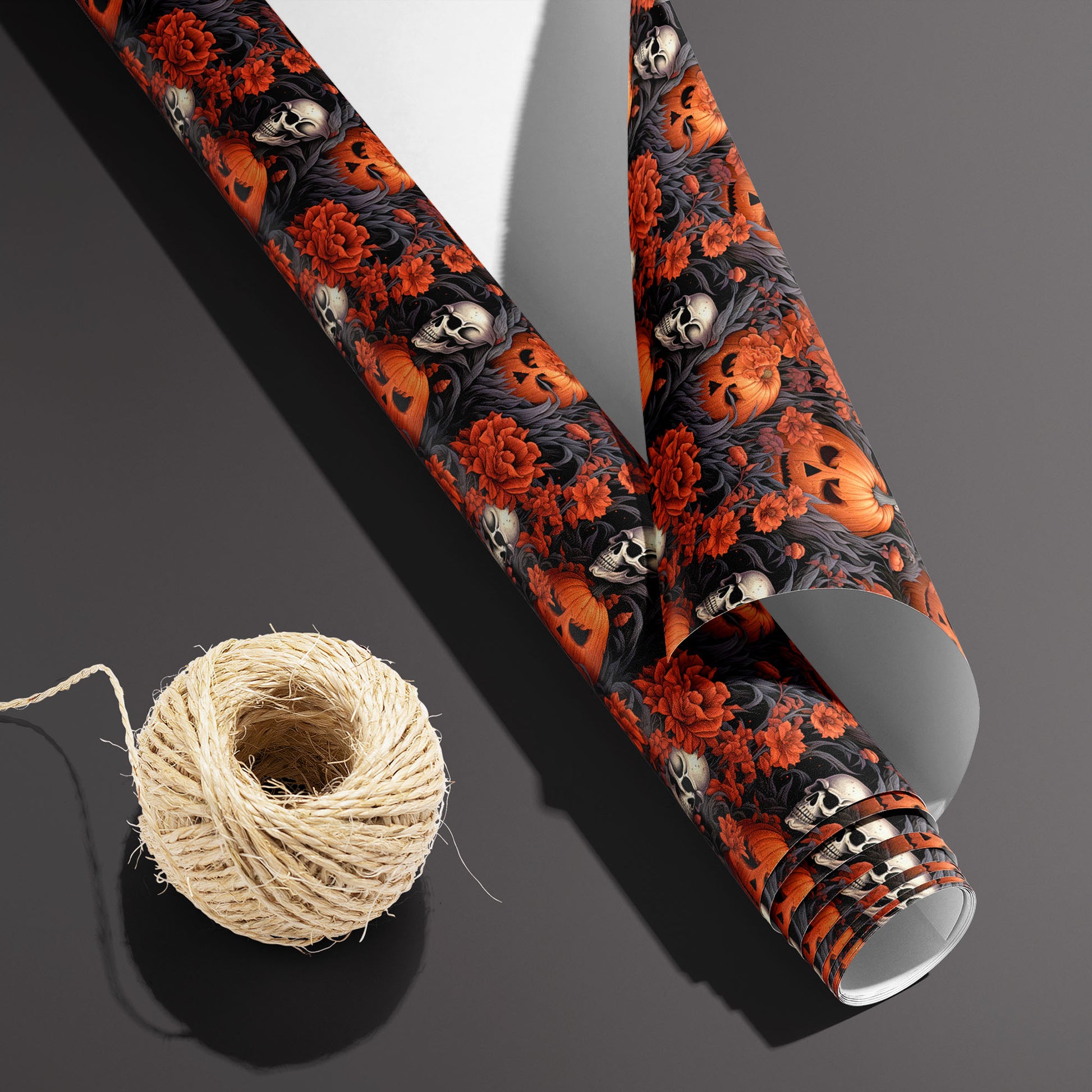 A partially unfurled roll of Ghastly Grove with the white underside visible beside a spool of natural twine, displaying pumpkins, skulls, and red blooms on black, on a dark gray surface.