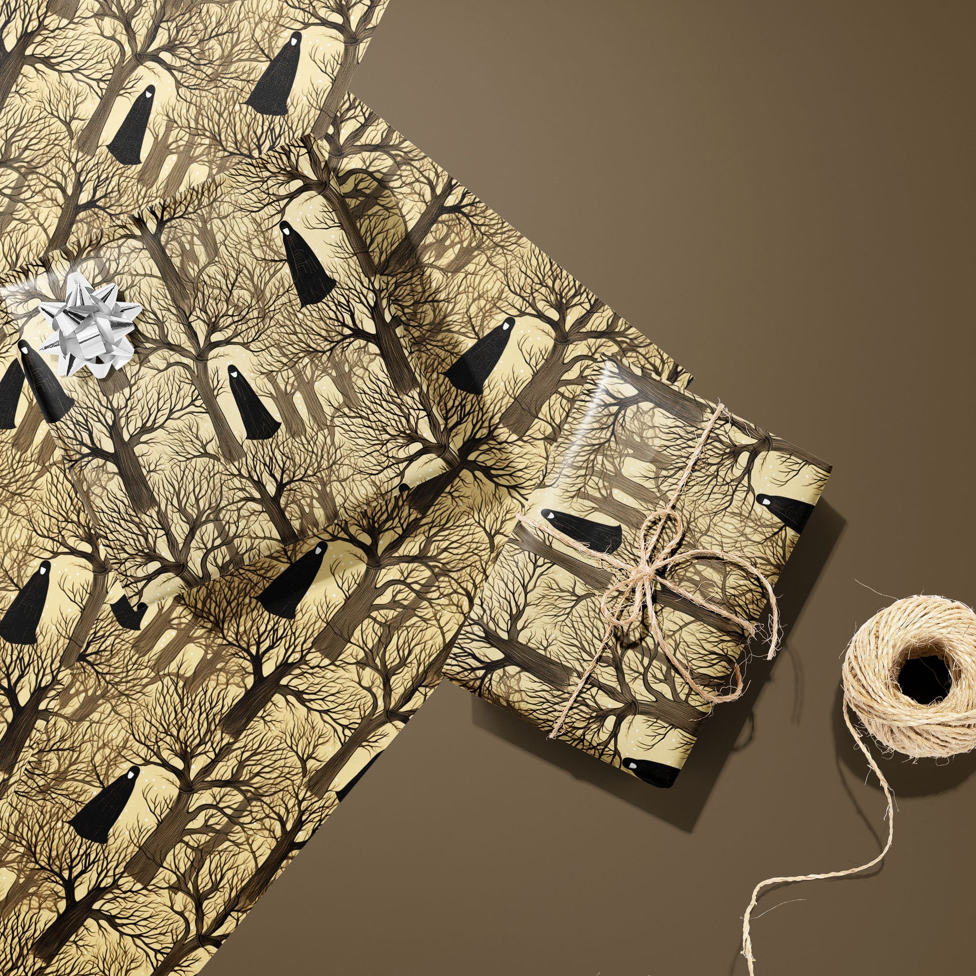 Two Silent Forest gifts—one with a silver bow and one tied with twine—rest on two unused sheets beside a twine spool on a warm brown surface, showing cloaked figures and bare trees.