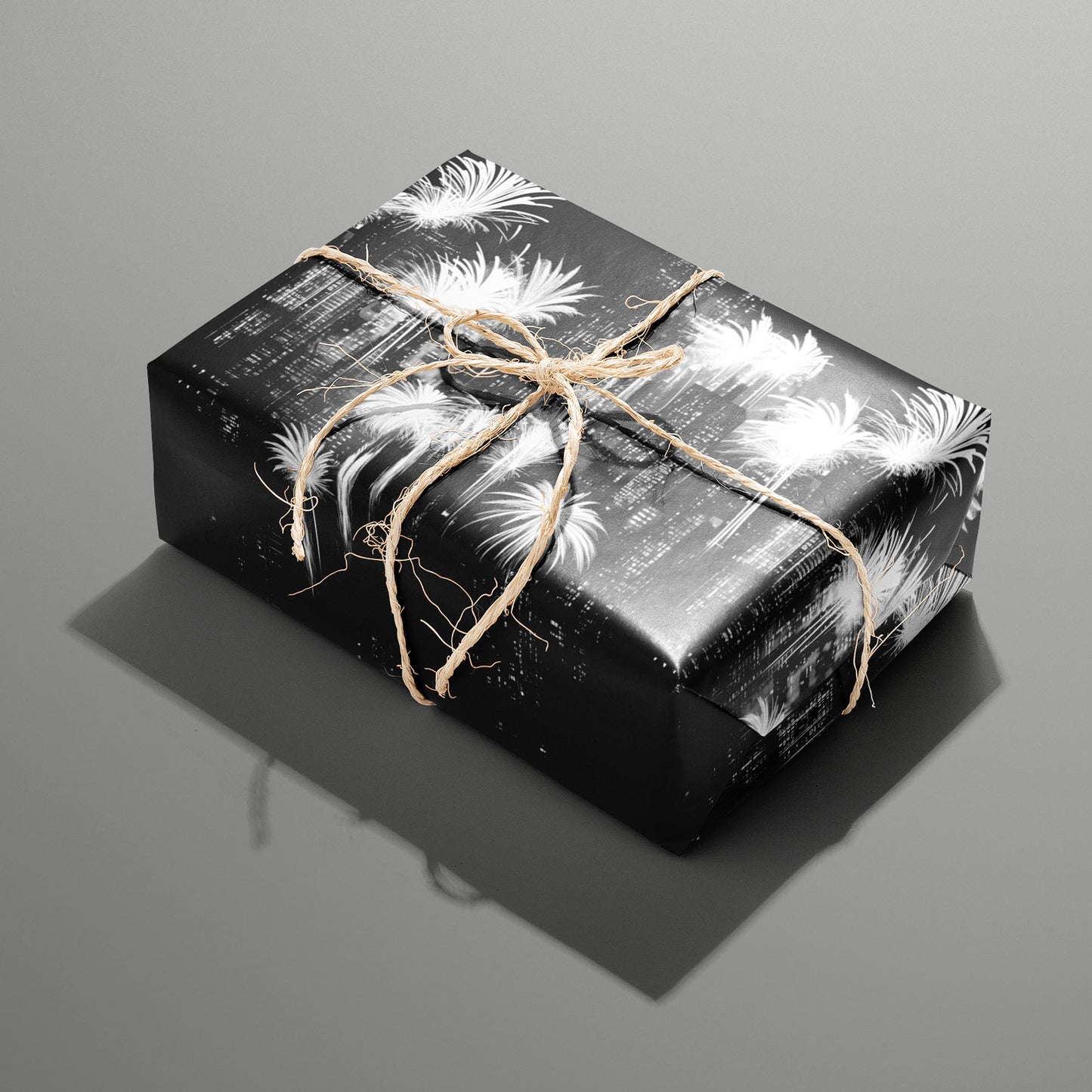 A single rectangular gift wrapped in Fireworks Fantasy paper—bright white fireworks bursts on black—tied with natural twine on a slate gray background.