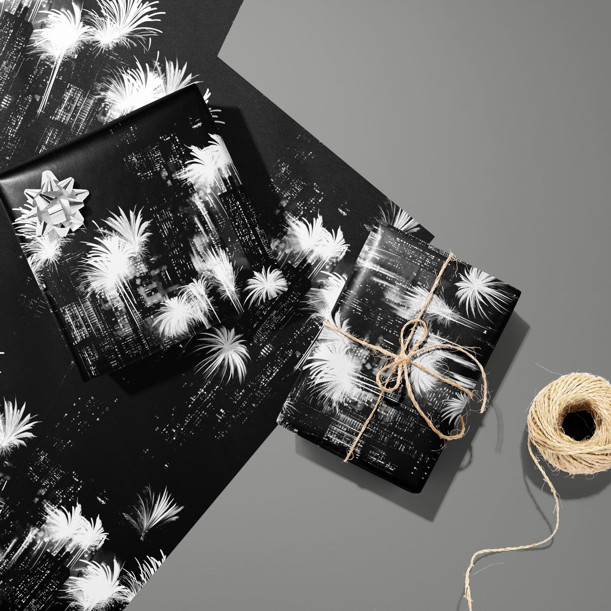 Two gifts in Fireworks Fantasy wrap—one with a silver bow and one tied with twine—rest on two unused sheets beside a spool of twine on a slate gray background.