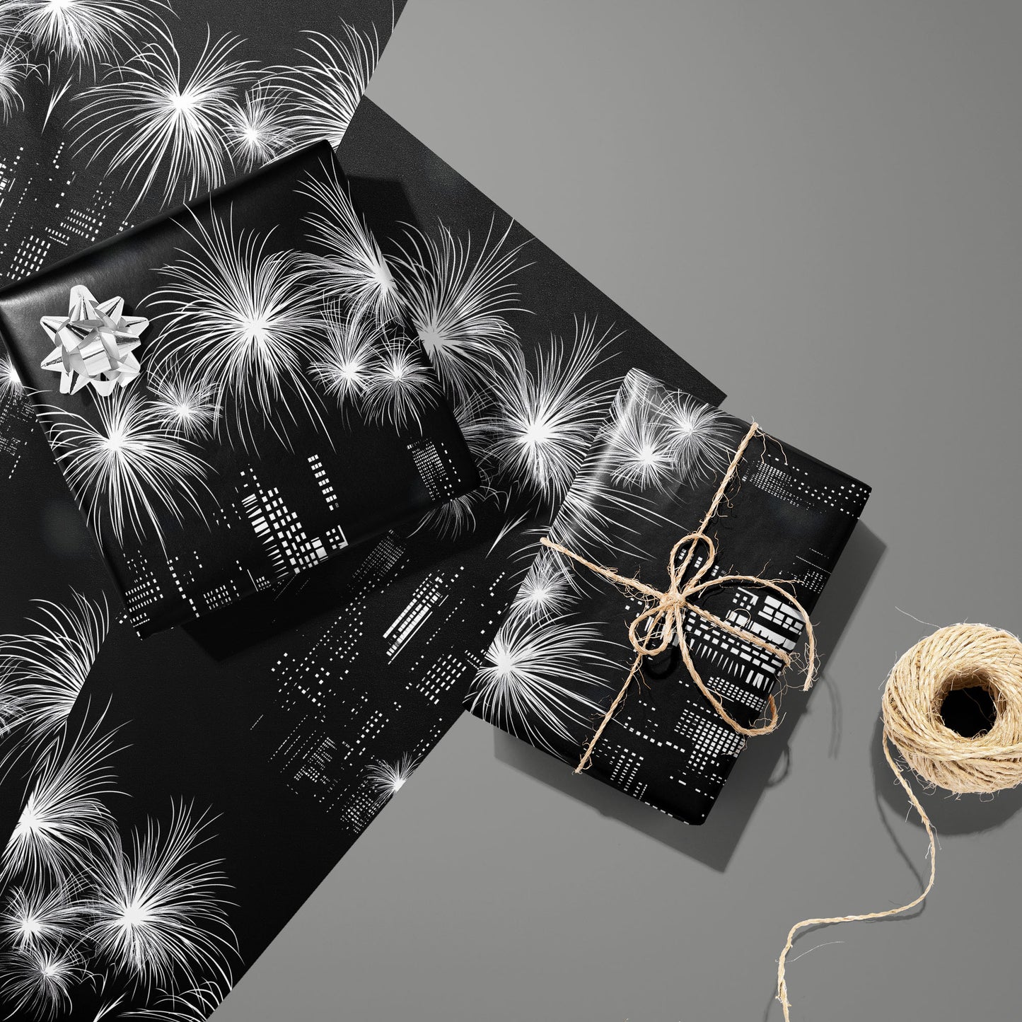Two gifts in Skyline Magic—one with a silver bow, one tied with twine—rest on two matching sheets beside a twine spool, showing a monochrome skyline and fireworks on a medium gray surface.