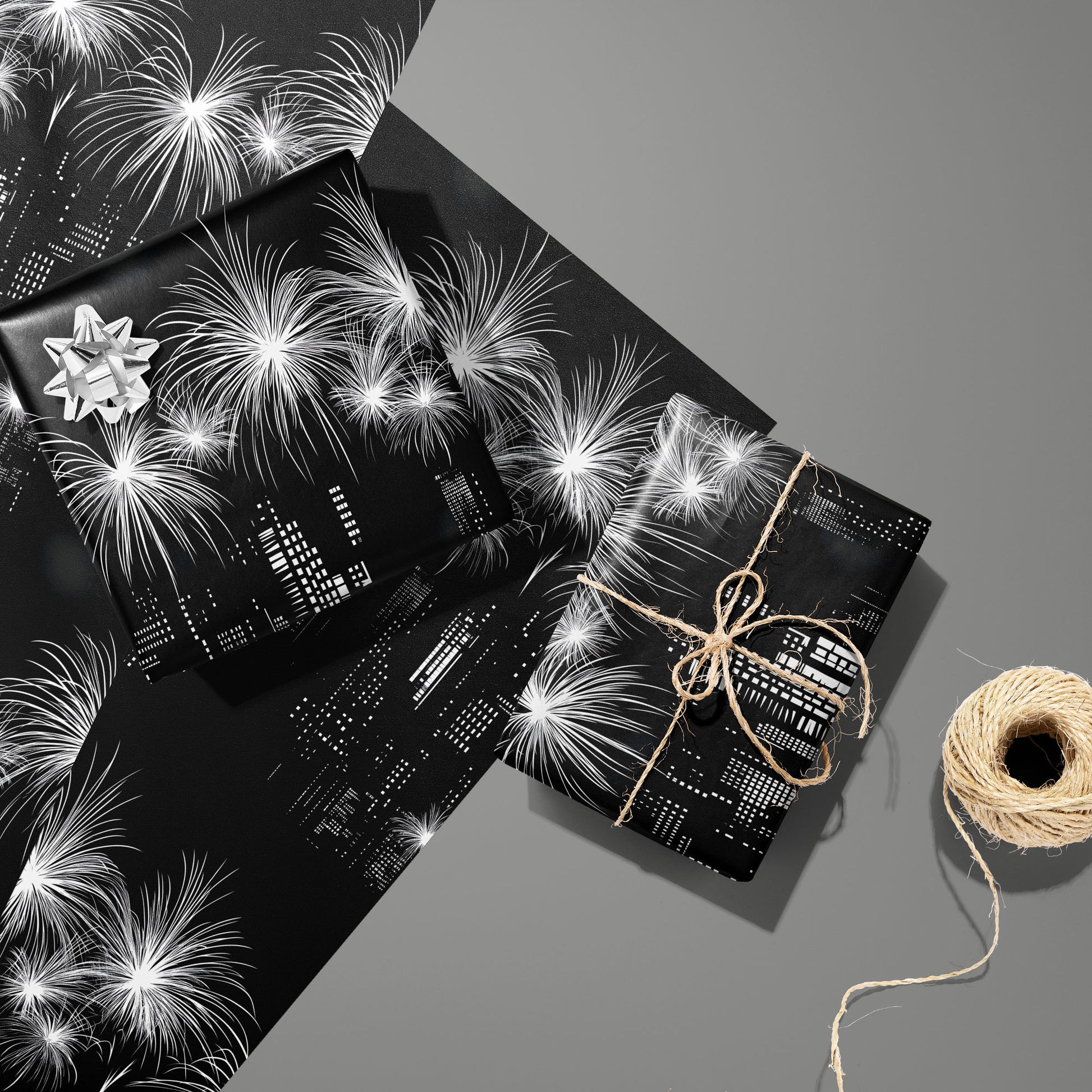 Two gifts in Skyline Magic—one with a silver bow, one tied with twine—rest on two matching sheets beside a twine spool, showing a monochrome skyline and fireworks on a medium gray surface.