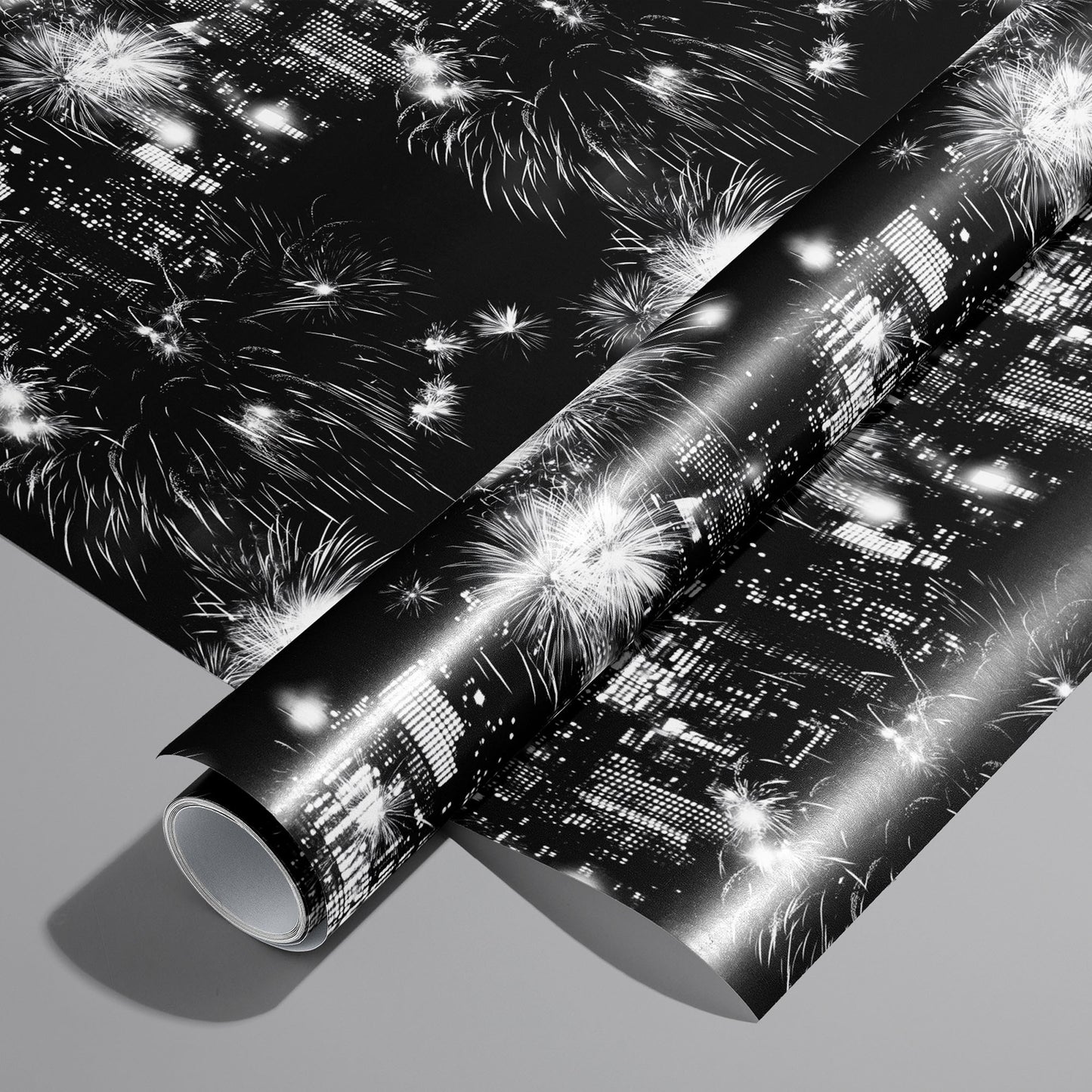 A roll on an unrolled sheet of Skyline Celebration wrapping paper with monochrome city skylines and bursting fireworks on black, set on a cool gray background.