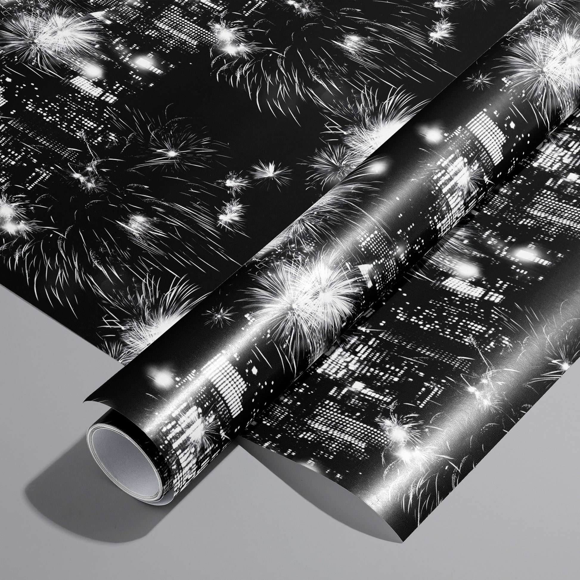 A roll on an unrolled sheet of Skyline Celebration wrapping paper with monochrome city skylines and bursting fireworks on black, set on a cool gray background.