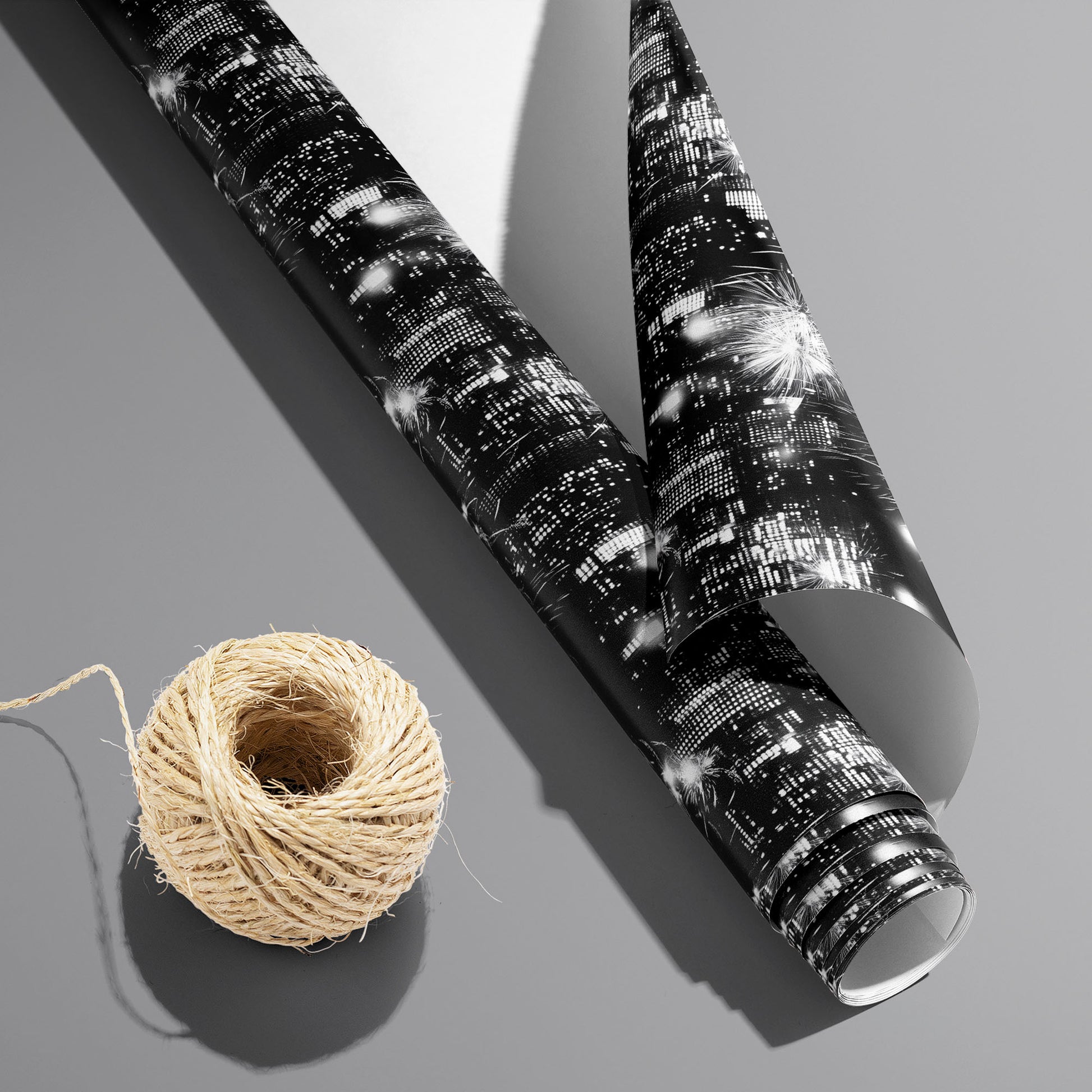 A partially unfurled Skyline Celebration roll with the white underside visible beside a spool of natural twine, showing city skylines and fireworks on a cool gray surface.