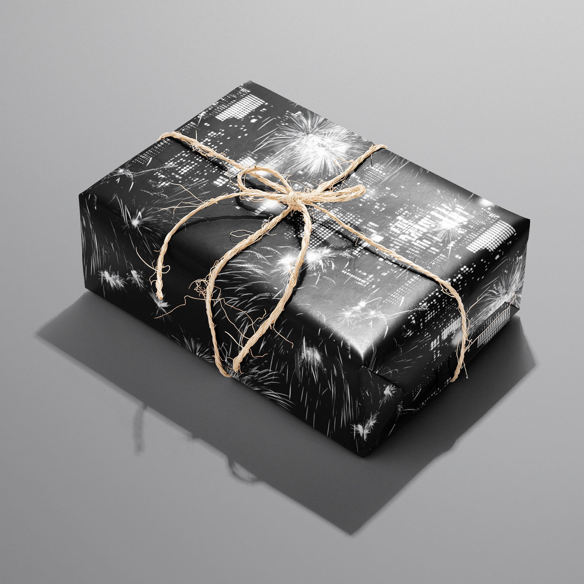 A single rectangular gift wrapped in Skyline Celebration—city lights and fireworks on black—tied with natural twine on a cool gray background.