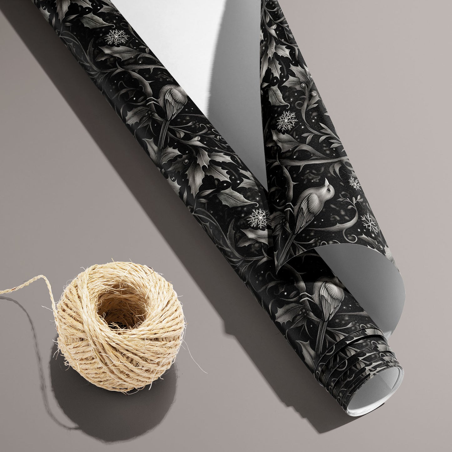 A partially unfurled roll of Winter Song with the white underside visible sits beside natural twine on a taupe surface, revealing birds, holly, and snowflakes on a black field.