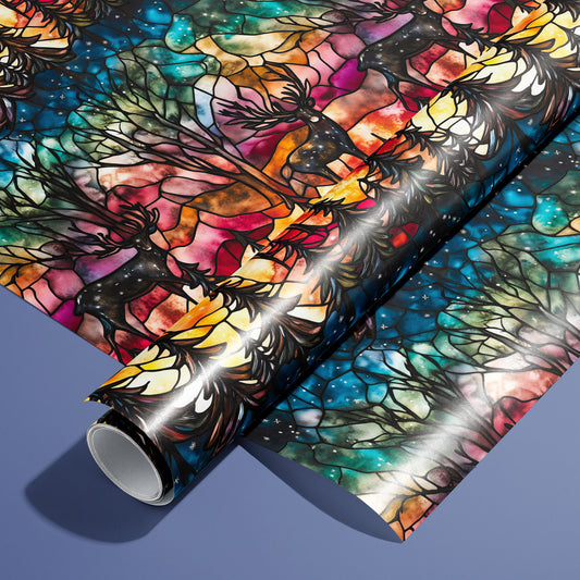 A roll of Starlit Grace wrapping paper on an unrolled sheet, featuring stained-glass woodland scenes with deer, fir trees, and starry skies in jewel tones on a slate blue surface.