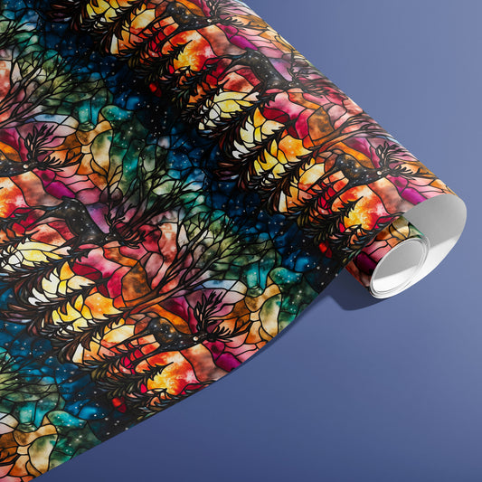 A partially unfurled roll of Starlit Grace wrapping paper showing stained-glass deer, fir silhouettes, and a star-dotted night in vivid aurora hues on a slate blue surface.