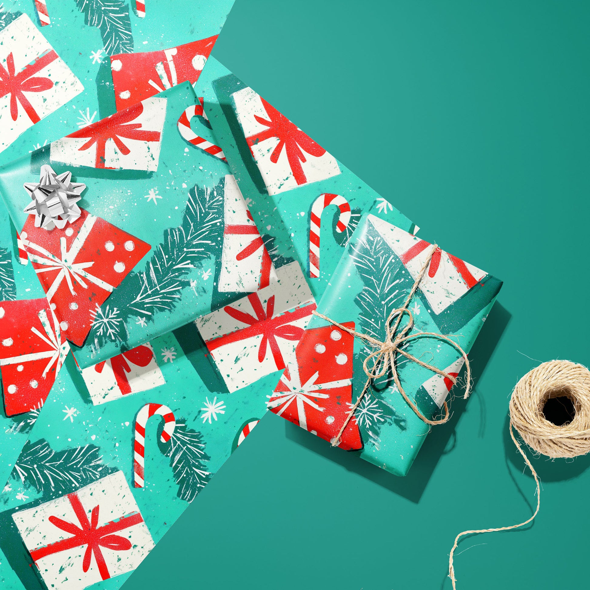 Two gifts in Peppermint Pop—one with a silver bow and one tied with twine—rest on two unused sheets featuring candy canes and pine sprigs beside a twine spool on teal green.