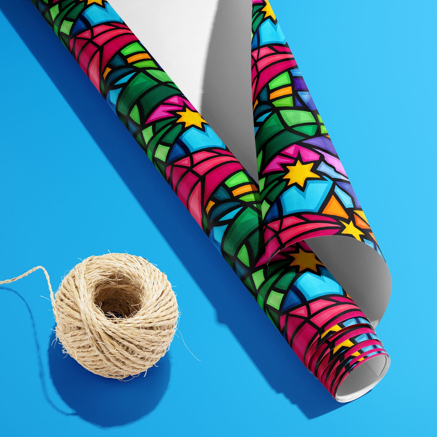 A partially unfurled Christmas Kaleidoscope roll with its white underside visible beside a spool of natural twine on a bright blue surface, featuring stained-glass stars and colorful facets.