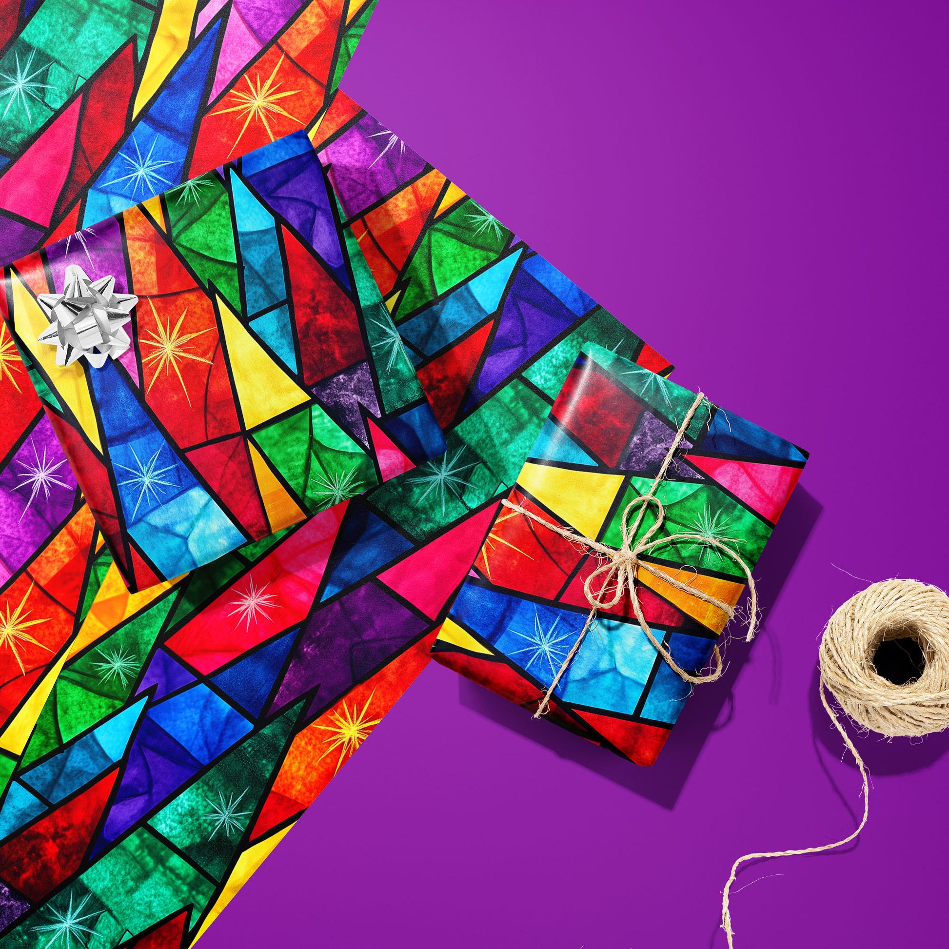 Two gifts wrapped in Prismatic Holiday—one silver-bowed, one tied with twine—rest on two unused sheets beside a twine spool on a vibrant purple surface with stained-glass starbursts.