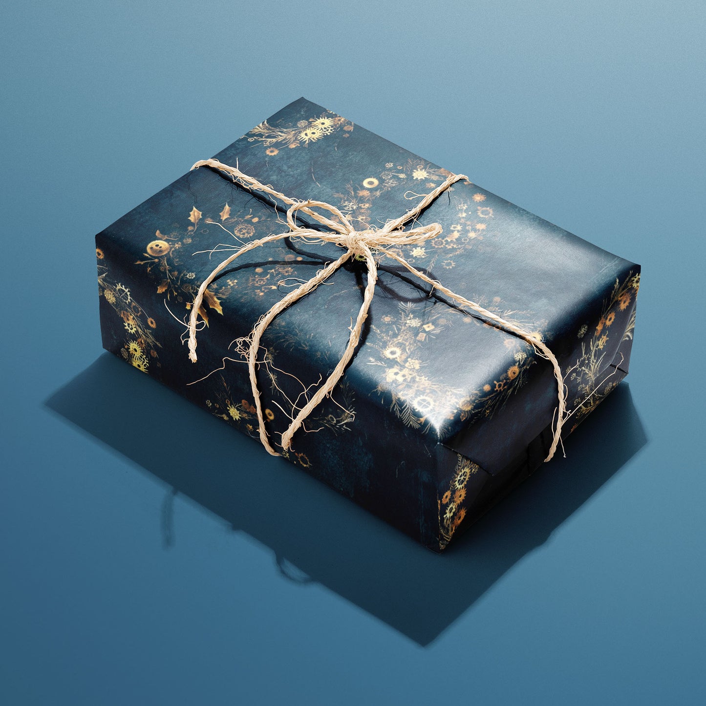 A single rectangular gift wrapped in Gilded Gears and tied with natural twine, the golden gear-and-holly wreaths glowing on a deep navy background.