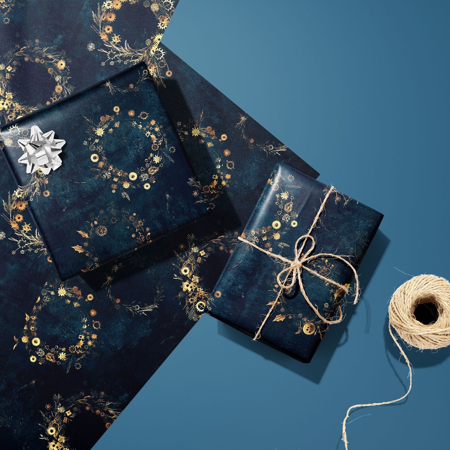 Two gifts in Gilded Gears—one with a silver bow, one tied with natural twine—rest on two unused sheets beside a spool of twine on a blue background, showing golden cog wreaths.