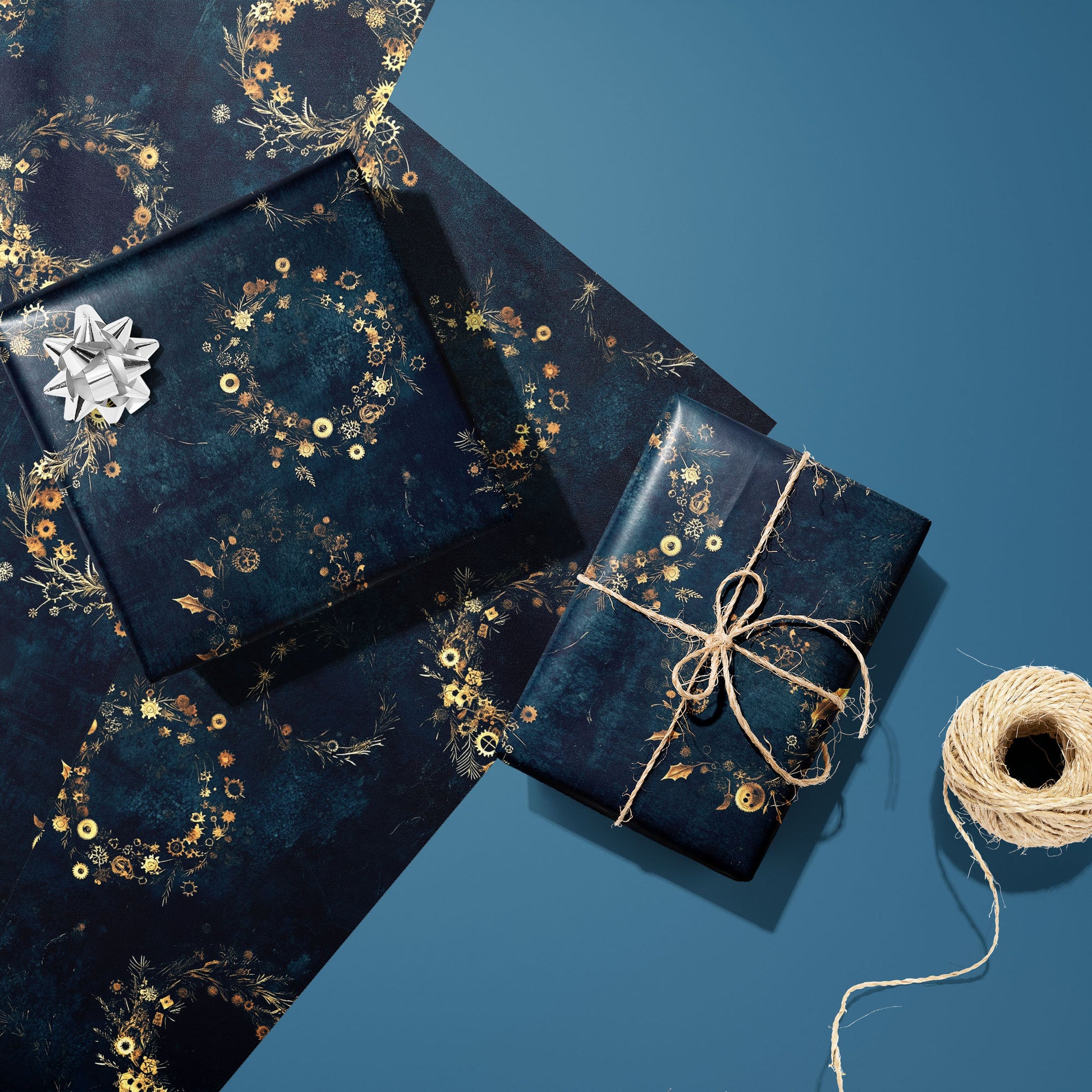 Two gifts in Gilded Gears—one with a silver bow, one tied with natural twine—rest on two unused sheets beside a spool of twine on a blue background, showing golden cog wreaths.