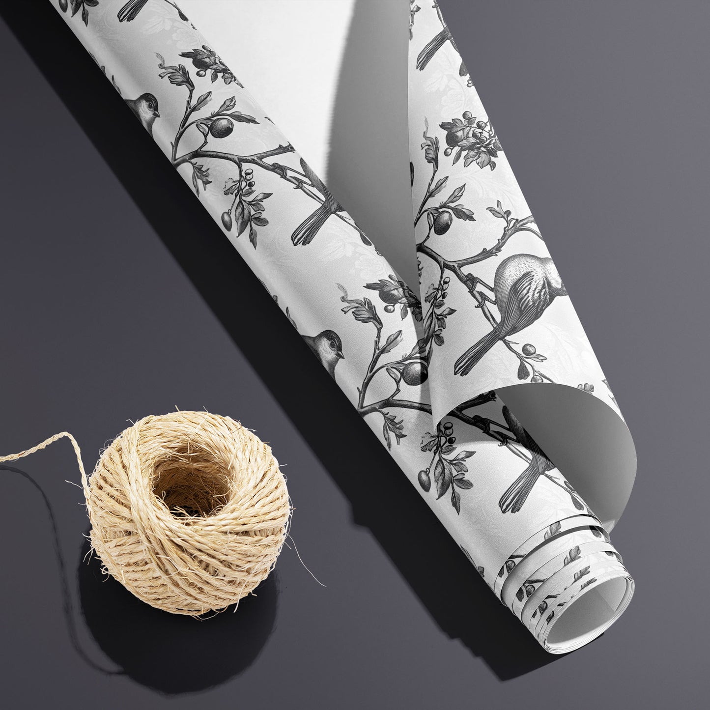 A partially unfurled roll of Winter Aviary, white underside visible beside a spool of natural twine, shows engraved birds on branching berries over a soft white damask ground on charcoal gray.