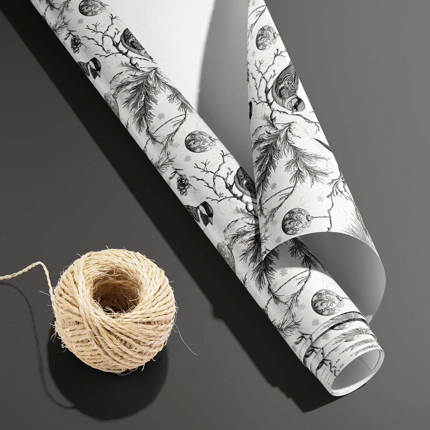 A partially unfurled Ornamental Nesting roll with its white underside beside a spool of natural twine on dark gray, displaying birds, pine boughs, cones, and ornaments on white.